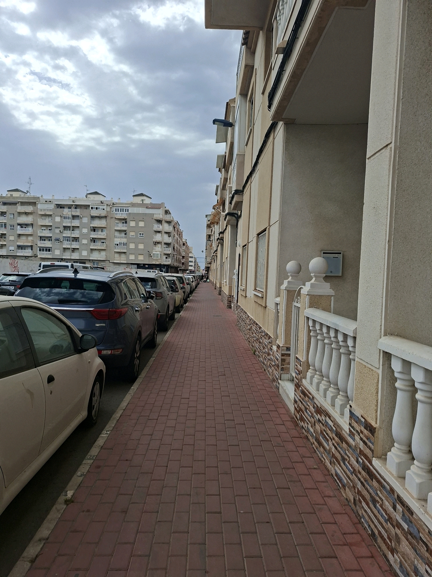 The image shows a street in Torrevieja, Spain, with parked cars lining the side of the road. A pedestrian sidewalk made of red brick pavers runs alongside a building. The building has a light-colored facade with multiple windows and balconies. The sky is overcast with gray clouds. The scene is one of an urban street during the daytime.