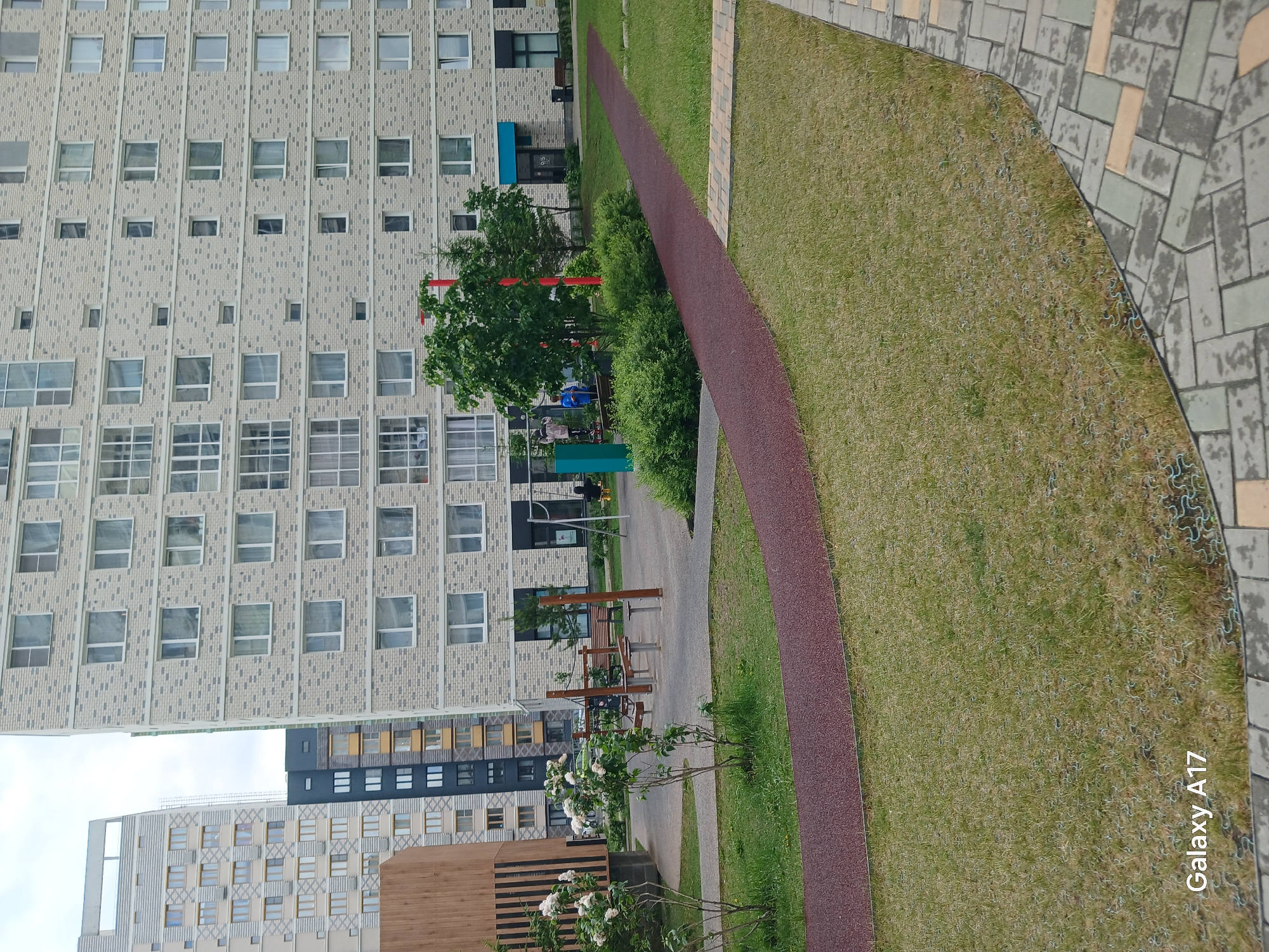 The image depicts an urban landscape in Yekaterinburg, Russia. Tall residential buildings with repetitive window patterns dominate the left side. A curved pedestrian path, bordered by green grass and concrete paving, runs diagonally from the bottom left to the top right. The path is made of reddish-brown material. A playground structure, visible between the buildings, features red and green elements. Some trees and shrubbery can be seen. A few people are present, though not clearly identifiable. A small wooden structure with white flowers stands in the foreground. The sky is overcast, and the lighting is somewhat muted. The photo includes the timestamp "Galaxy A17".