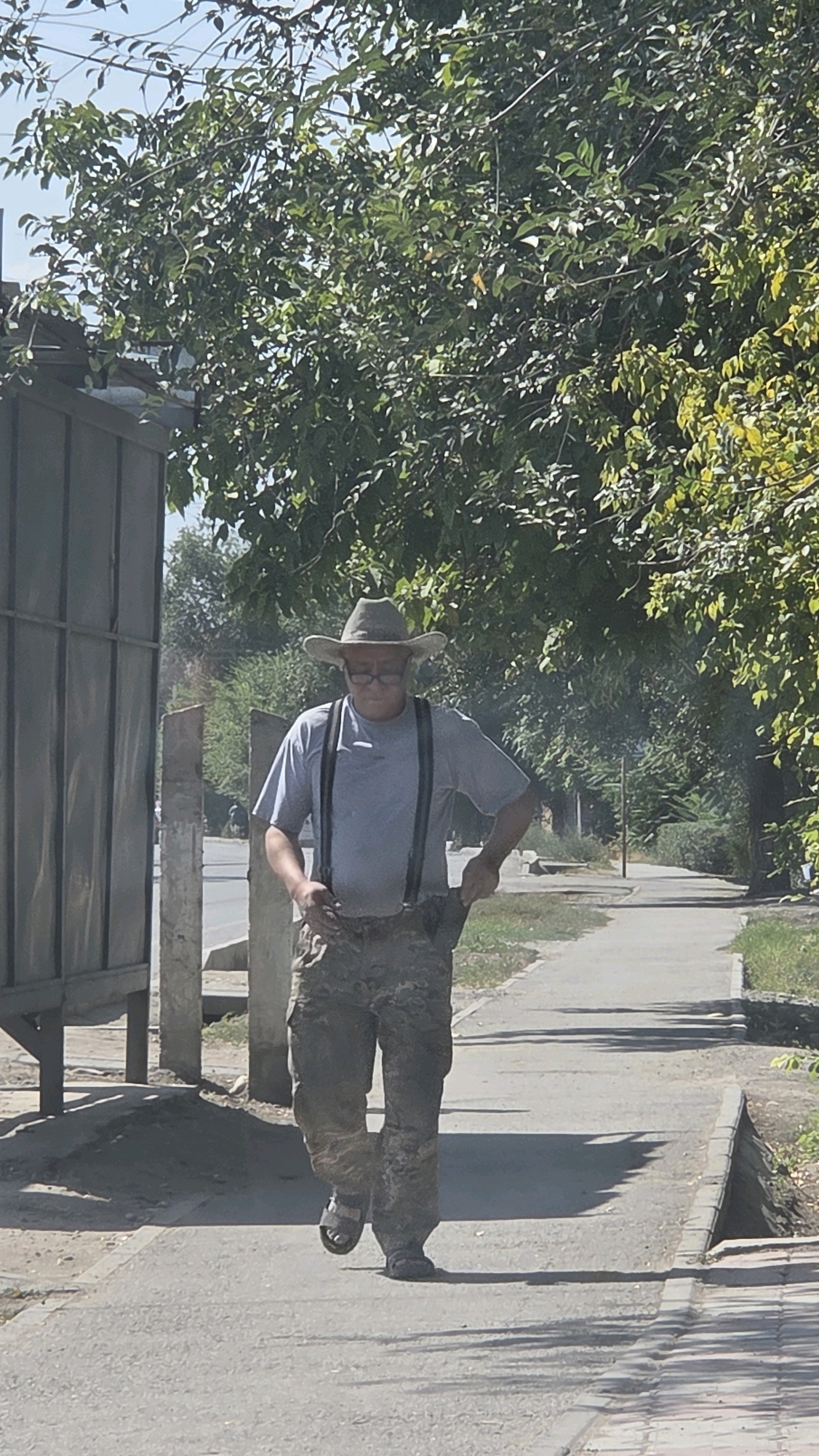 An adult male wearing a light-colored cowboy hat, glasses, a grey t-shirt, dark suspenders, and camouflage pants walks along a paved sidewalk. He is also wearing dark sandals. He appears to be reaching into his right pants pocket. Behind him, a metal fence runs along the left edge of the image, and behind the fence, a street with some traffic is visible. The sidewalk is bordered by a grass area and trees. The sky is visible above the trees. The scene is set in Tokmok City, Kyrgyzstan, on a sunny day.