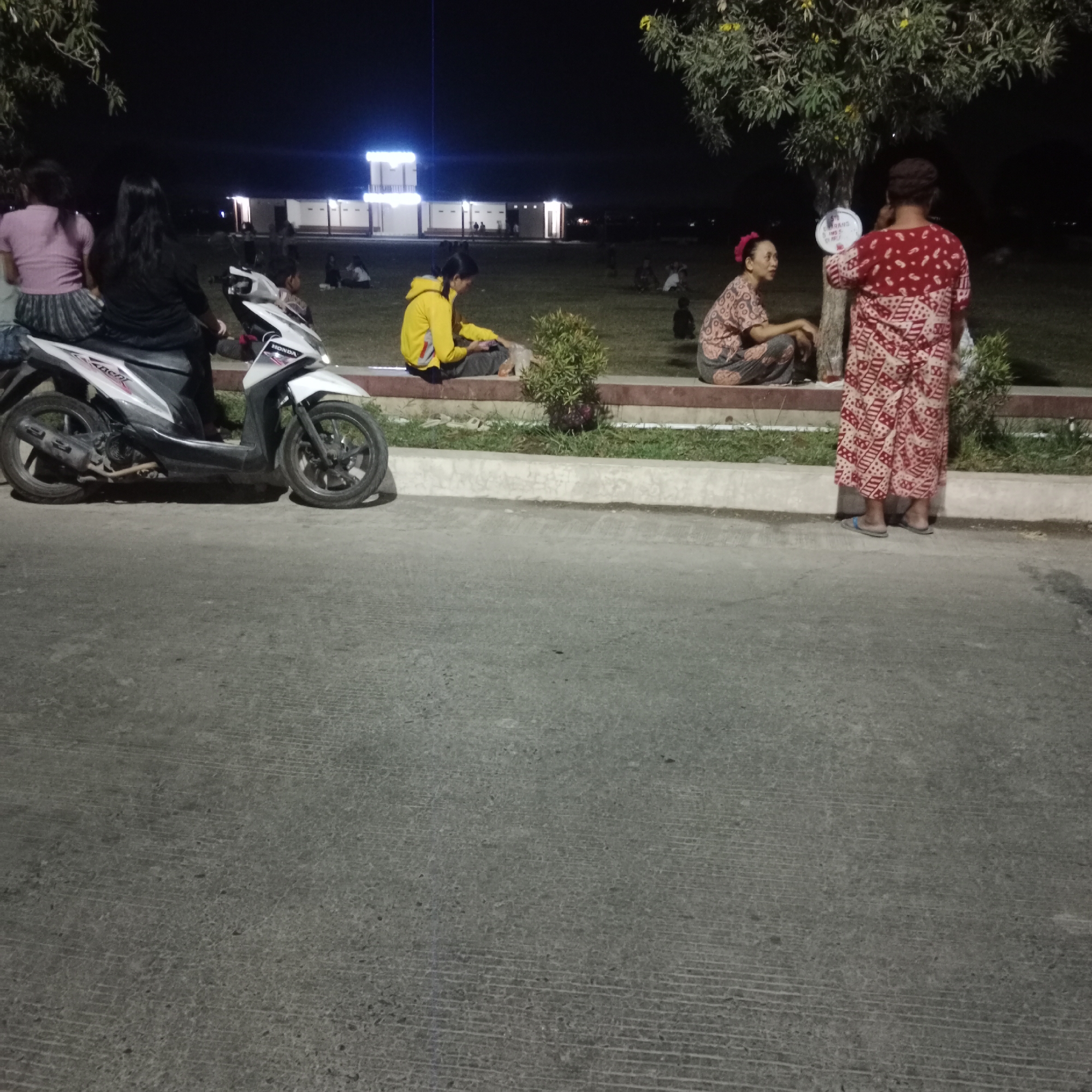 The image depicts a nighttime scene in what appears to be a public park or gathering space in Kunjeng, Indonesia. In the foreground, a white Honda Beat motorcycle is parked on a paved surface. To the left of the motorcycle, two women are seated, one in a pink top and the other in a black top, with their backs to the camera.
Further back, on a raised grassy area bordered by a concrete and brick ledge, several other individuals are present. A person in a yellow hoodie is seated on the ledge, looking at a phone. To their right, a woman in a patterned pink and brown outfit is also seated. Standing near a tree is a person wearing a red and white patterned garment, holding a circular object, possibly a fan, and facing another seated woman.
In the background, there are low buildings with illuminated windows, suggesting a community area or marketplace. Trees with sparse foliage are visible, casting shadows in the low light. The overall atmosphere is relaxed, with people gathered outdoors in the evening. The presence of the motorcycle and the general setting suggest a casual community event or a place where people congregate.
