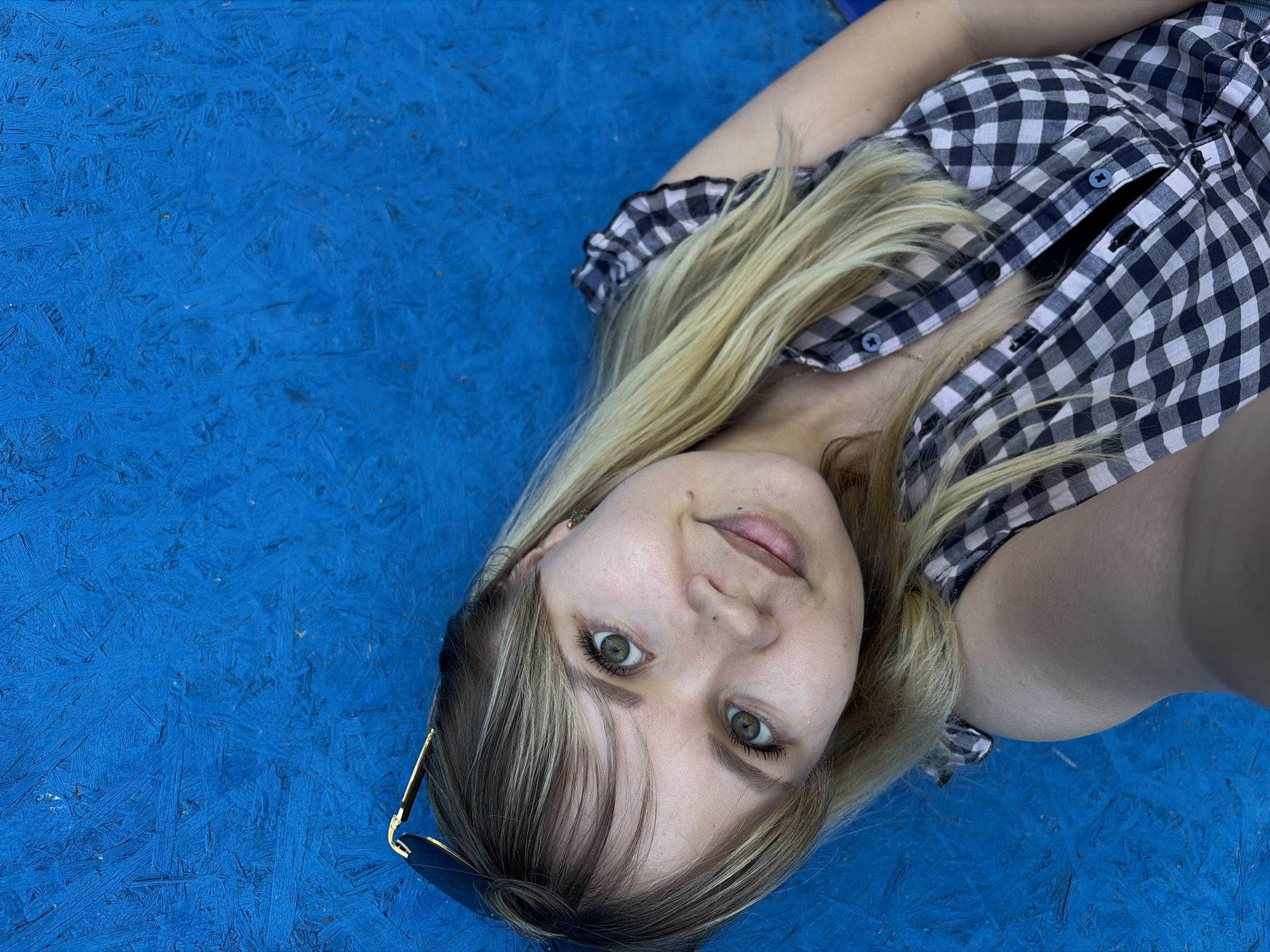 A young woman lies on a blue textured surface. She has blonde hair with dark roots, fair skin, and green eyes. She wears a black and white checkered button-up shirt, partially unbuttoned. Sunglasses are placed near her face. The location is Чумна, Russia.