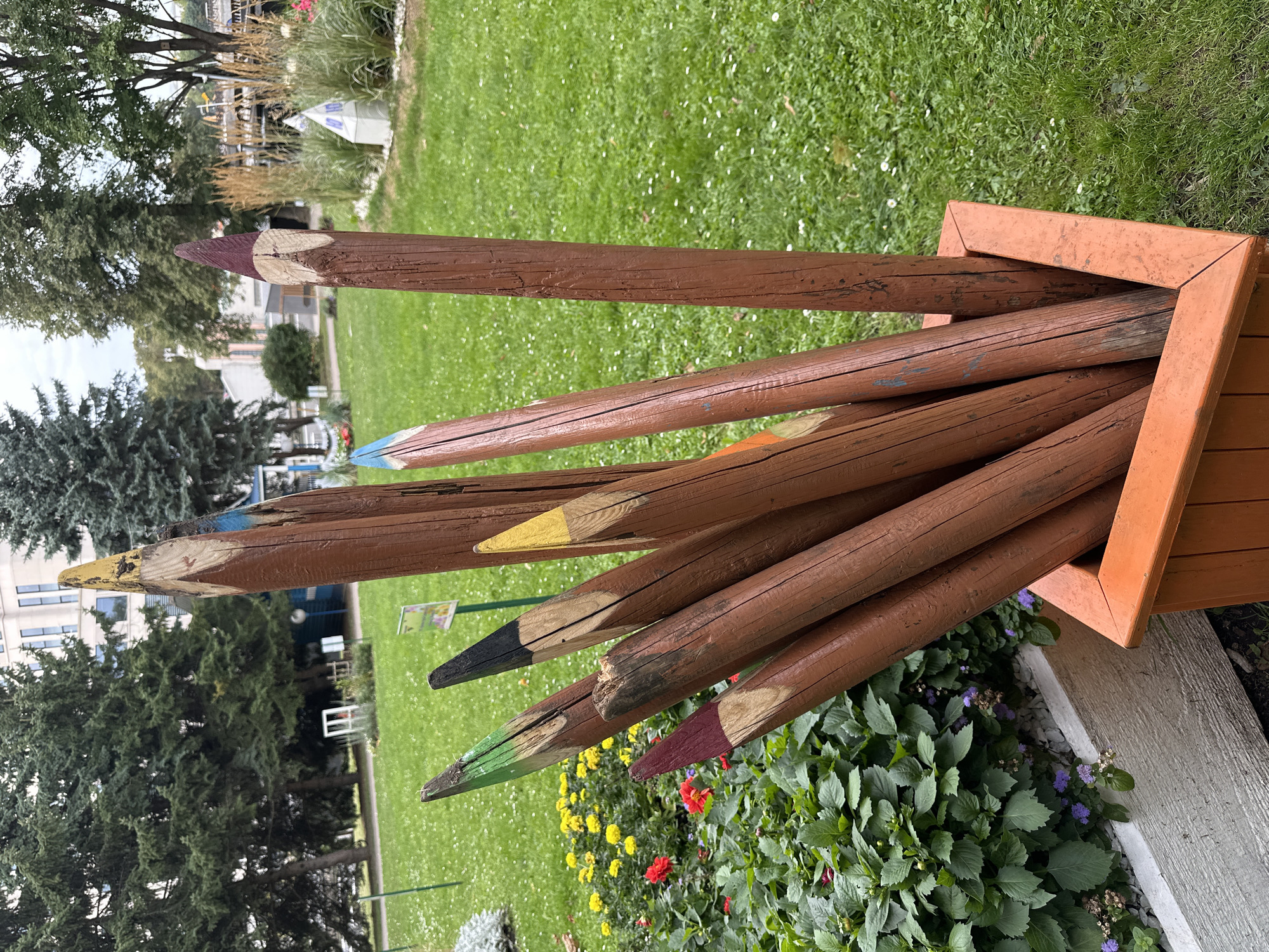 An outdoor park scene in Minsk, Belarus, features a cluster of oversized, wooden pencils standing upright in an orange planter box. The pencils are painted in various colors at their tips, mimicking the appearance of sharpened colored pencils. Some of the pencil tips are painted green, black, yellow, maroon, and blue. The wooden shafts are a reddish-brown color and show signs of weathering.
Behind the planter, a lush green lawn is dotted with small white flowers. To the left of the planter, a raised flower bed contains green foliage and various blooming flowers, including yellow and red blossoms. In the background, several mature evergreen trees provide a backdrop, with hints of buildings and other park structures visible through the branches. The sky appears overcast. There are no people visible in the frame.