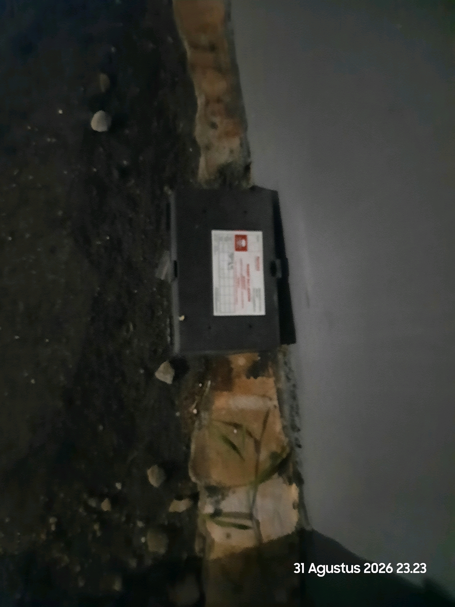 A black utility box with a white label is situated on the edge of a paved surface next to a dirt area. The paved surface is a light gray, and the dirt area is dark and uneven, with small stones scattered throughout. A textured, rough surface, possibly brick or concrete, runs along the border between the pavement and the dirt. Some dry vegetation is visible near the utility box. The image is dated "31 Agustus 2026 23.23". The location is Gending, Indonesia.