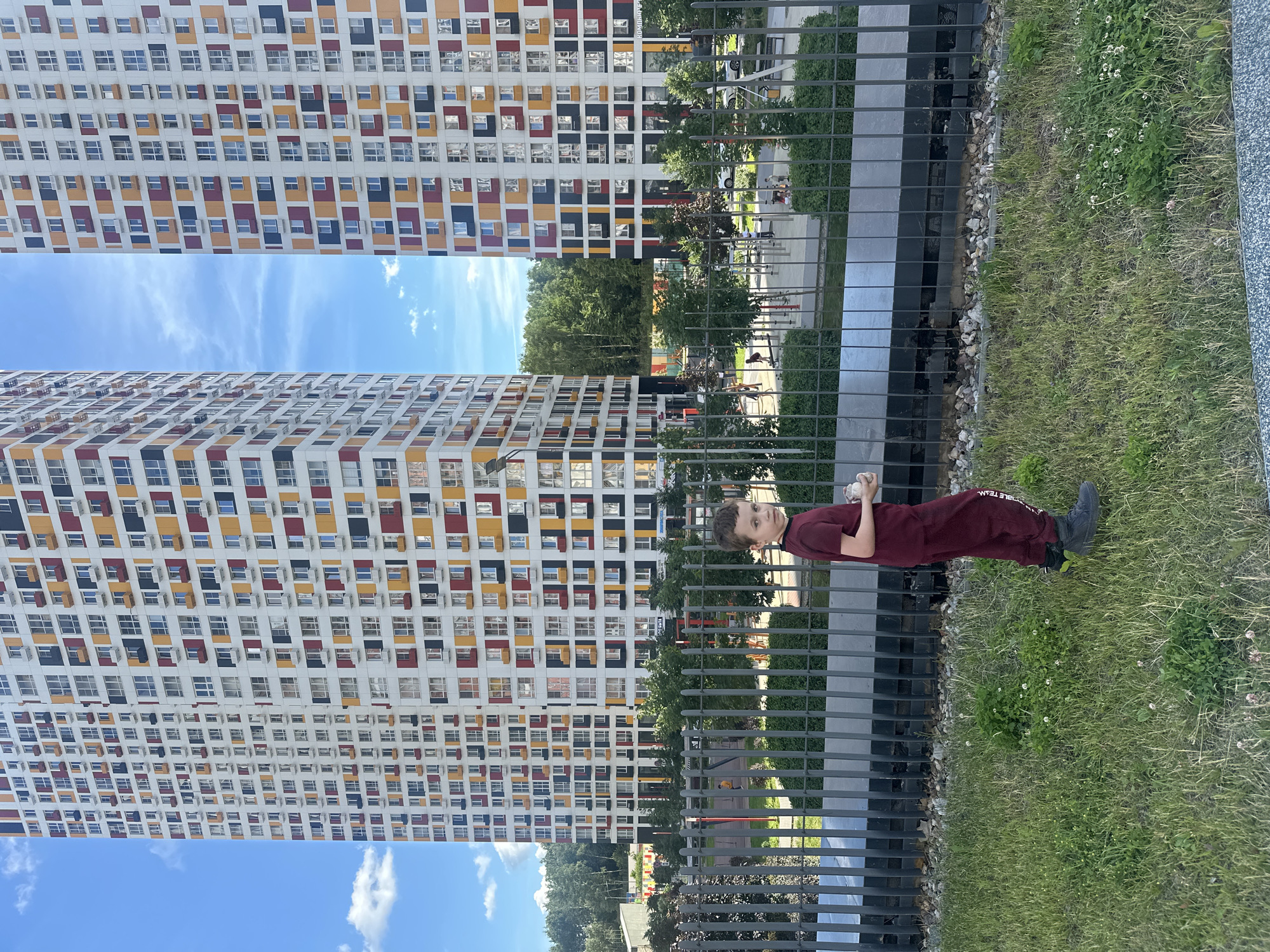 A young boy stands in the foreground, facing the camera with a slight turn to his right. He is wearing a maroon t-shirt and matching maroon sweatpants, with dark gray shoes. He is holding a small, light-colored object in his right hand. The boy is positioned in front of a metal fence, behind which is a playground area with swings and play structures. In the background, two towering, modern apartment buildings stand tall, their facades adorned with a colorful pattern of red, yellow, and dark blue panels interspersed with white and windows. The sky above is a bright blue with scattered white clouds. The environment appears to be an urban residential area on a clear day.