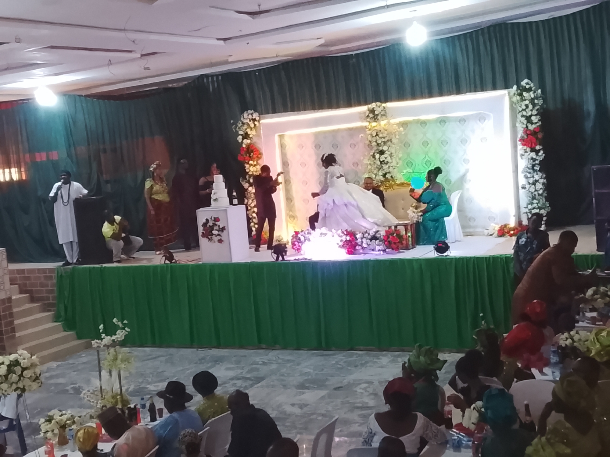 The image shows a wedding reception in Jos, Nigeria. The bride, in a white gown, and groom are on a stage decorated with flowers and white fabric. A tiered wedding cake is visible on the left side of the stage. Several people are on the stage, including a person speaking at a podium, and others interacting with the couple. Guests are seated at tables in the foreground. Green fabric drapes the front of the stage. The lighting is provided by overhead fixtures.