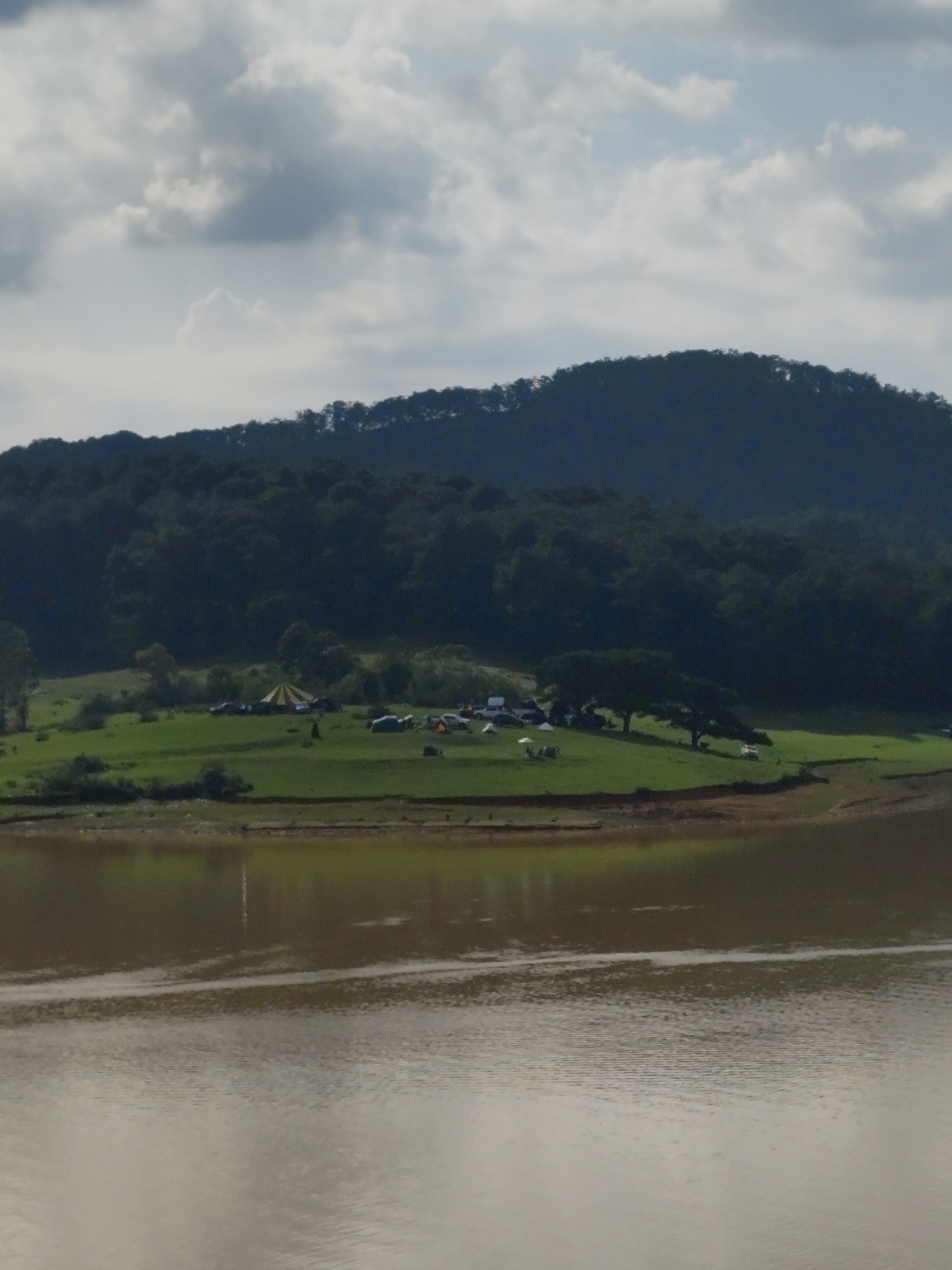The image presents a scenic outdoor landscape in Lạc Dương, Vietnam, featuring a prominent body of water in the foreground, likely a lake or reservoir, with its surface showing subtle ripples and reflecting the overcast sky. The water itself appears a muddy, brownish-grey color.
Beyond the water, a vibrant green grassy area stretches across the middle ground, gently sloping towards a dense forest. This grassy expanse is the setting for an active outdoor gathering, possibly a camping event or festival. Numerous tents are pitched across the field, with a large, distinctive yellow and white striped tent standing out centrally. Smaller individual camping tents in various colors like blue, grey, and white are also visible. Several vehicles, including cars or SUVs, are parked near the tents, indicating accessibility to the site. A large, leafy tree provides ample shade to a section of the campers, and at least one white umbrella is also set up.
The background is dominated by a majestic, dark green mountain range, thickly covered in trees, extending across the horizon. The sky above is overcast, filled with a mix of light and darker grey clouds, suggesting a cloudy day but with sufficient natural light. The scene conveys a sense of peaceful natural beauty combined with community and outdoor leisure activities. No visible text is present in the image.