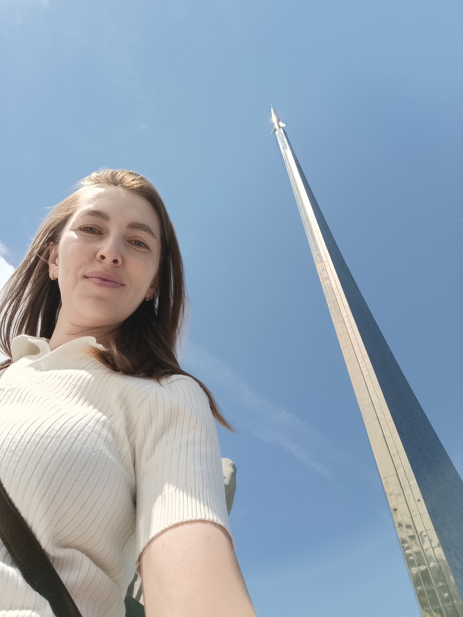 An outdoor, low-angle shot captures a young woman in the foreground with a prominent monument in the background, set against a bright, clear blue sky. The woman, with long brown hair, occupies the left side of the frame, looking directly towards the viewer with a neutral to slightly smiling expression. She is wearing a light-colored, ribbed short-sleeved top, and a dark strap from a bag is visible over her left shoulder.
In the right background, a towering, slender metallic monument, identifiable as the Monument to the Conquerors of Space in Moscow, Russia, rises sharply towards the sky. The monument has a reflective surface and tapers to a point, topped by a small, distinct rocket model. Its imposing height and sleek design dominate the right side of the frame, emphasizing the upward perspective of the photograph. The sky is predominantly clear, indicating a sunny day with fine weather. There is no visible text in the image.