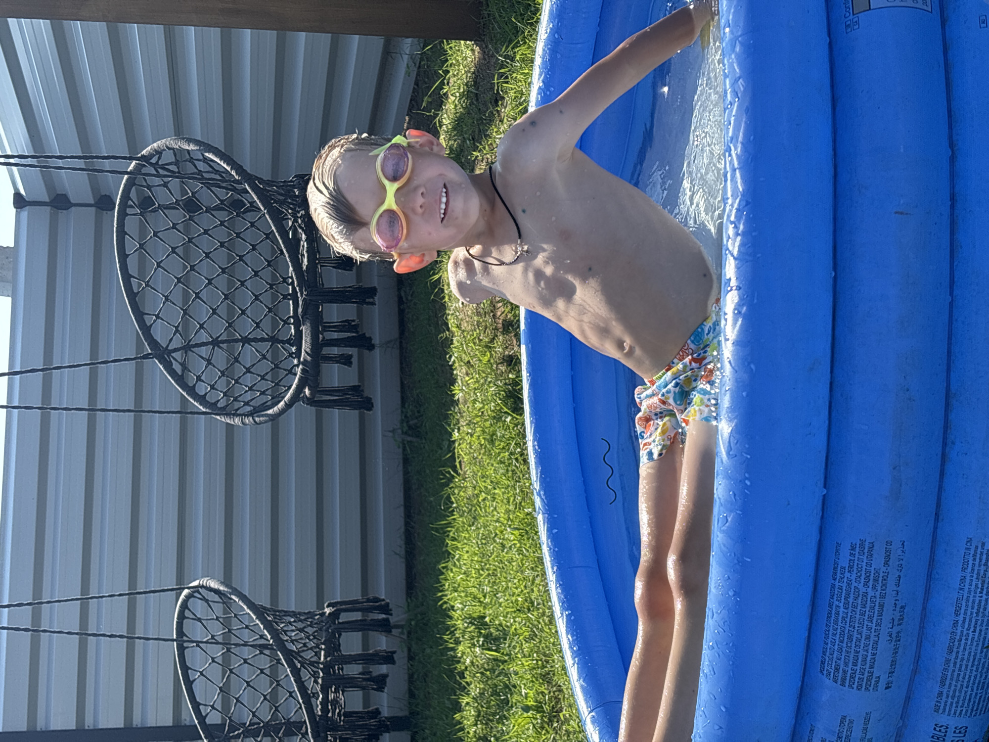 This is a close-up, slightly angled shot of a young boy in an inflatable pool on a sunny day. The boy, wearing yellow-rimmed goggles with orange ear pieces and patterned swim trunks, is smiling and looking at the camera. He is partially submerged in the blue pool, with water splashing around him. The pool is set on a patch of green grass, with a white corrugated metal structure and two dark, woven hanging chairs visible in the background. The lighting suggests it is daytime, likely a warm, sunny afternoon. The text visible on the pool's side appears to be a warning about drowning and manufacturing information, indicating the pool is made in China.