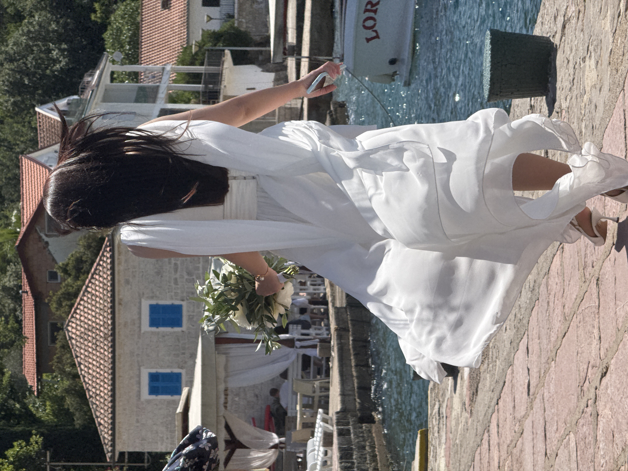 The scene depicts a woman in a white, flowing gown walking on a paved promenade alongside a body of water under bright sunlight. The location is in the city of Unknown, Unknown. The woman has long, dark brown hair that appears to be flowing or windblown, obscuring her face from the back. She is wearing white high-heeled shoes. In her left hand, she holds a white floral bouquet with prominent green foliage, and a delicate gold-colored bracelet is visible on her wrist. Her right hand holds a light-colored smartphone.
To the woman's right, calm blue water with sparkling reflections indicates a sunny day, with a white boat partially visible, bearing red letters that spell "LOR". A dark green, ribbed cylindrical object or pot rests on the paved ground near the water's edge. The promenade is constructed of reddish-brown or terracotta-colored paving stones.
In the background, traditional-style buildings with red-tiled roofs are visible. One prominent light-colored building directly behind the woman features two windows with bright blue shutters. Further back, more buildings with white facades and balconies are visible on a slight incline, surrounded by lush green trees and foliage. Near the water's edge, under white awnings, outdoor seating areas with tables and chairs are set up, with blurred figures of people seated.