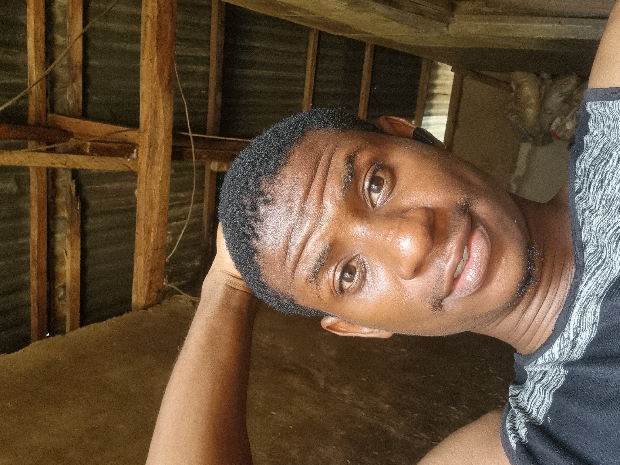 This media file is a selfie portrait of a young Black man, captured with the image rotated 90 degrees clockwise. The man, appearing to be in his 20s or early 30s, looks directly at the camera with a calm expression and a faint, engaging smile. He has short, dark, curly hair, brown eyes, a subtle mustache, and a short goatee. His skin is smooth and appears slightly reflective, possibly indicative of warmth or humidity, aligning with the provided context of Onitsha, Nigeria. He is wearing a dark-colored t-shirt with a distinctive light-colored, wavy or striped pattern visible on the shoulder and sleeve.
The setting appears to be an indoor space or a covered outdoor structure, characterized by rustic wooden beams and planks forming a visible support or framework. Behind these wooden elements, dark, corrugated metal sheets are visible, suggesting a basic or utilitarian construction. The floor beneath him appears to be bare earth or unfinished concrete. Thin wires are visible running across some of the wooden structures. In the upper right background, some light-colored, bundled objects, possibly plastic bags or discarded items, are faintly visible and partially obscured. The lighting in the image suggests it was taken during the daytime. The man's posture, with one arm bent and resting behind his head, conveys a relaxed and casual mood. No visible text is present in the image.