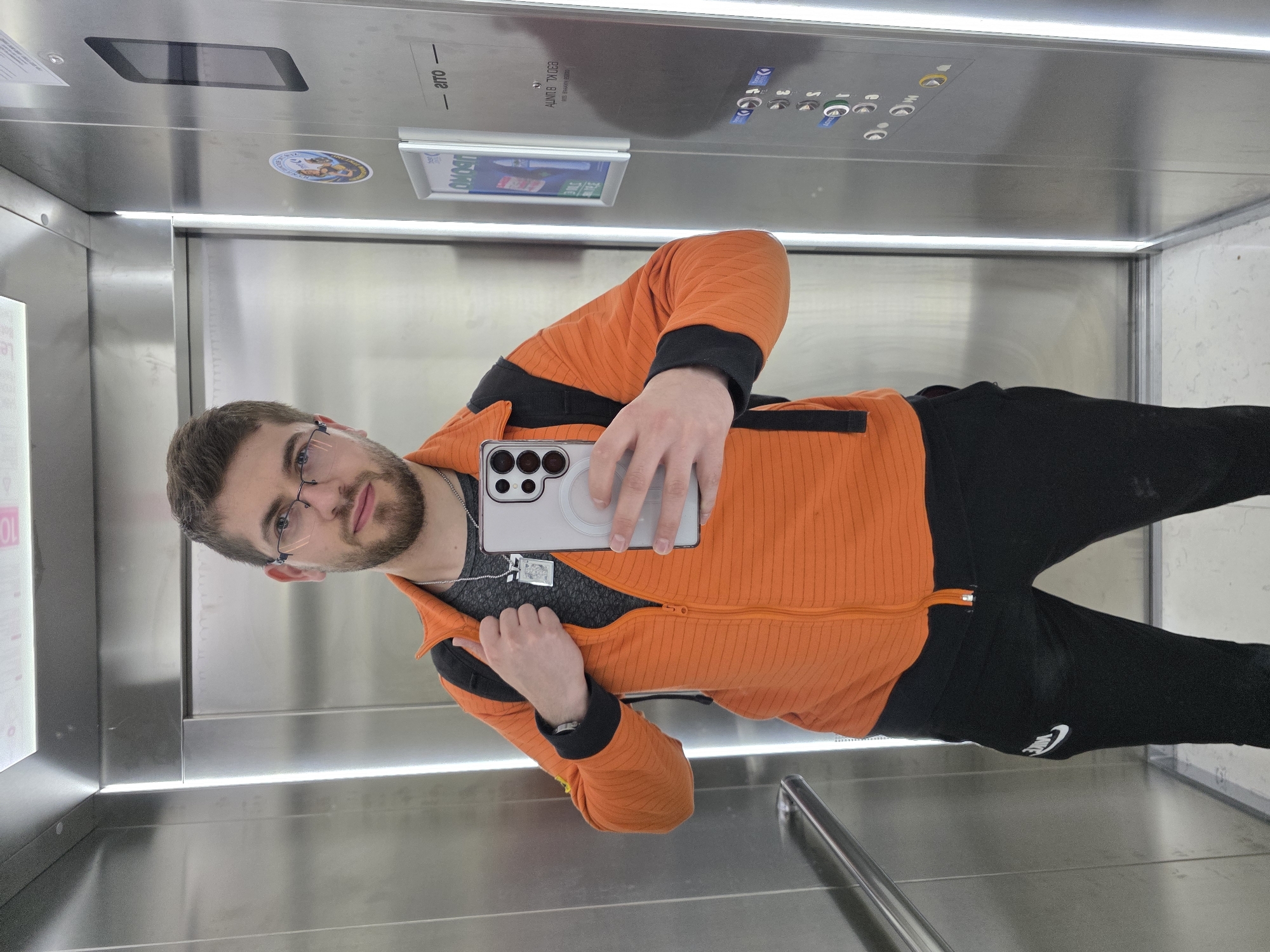 A man is taking a selfie in an elevator. He is wearing glasses, an orange jacket with black accents, a gray shirt, black pants with a Nike logo, and a silver chain. The elevator has stainless steel walls, buttons on the top, and a display. The image was taken in Plovdiv, Bulgaria.