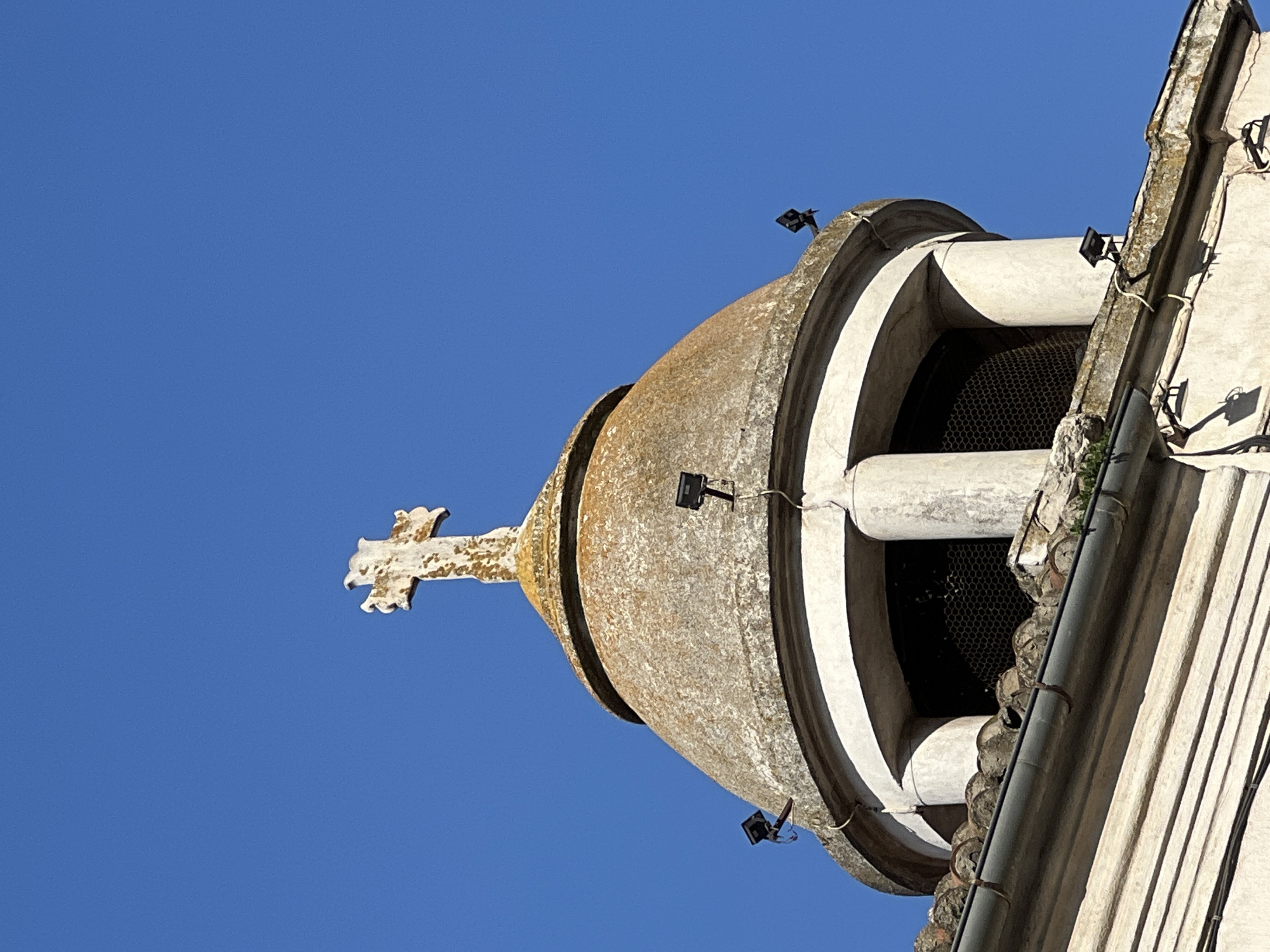 The image shows a close-up, low-angle view of the top of a building, likely a church, against a clear blue sky. A weathered, stone dome is visible, topped with a cross that also appears to be made of stone and shows signs of age and lichen growth. There are three small, black floodlights attached to the dome, each with visible wiring. The dome is supported by white, fluted columns that frame a dark opening, possibly a window, covered with a wire mesh. Below the dome, a section of a tiled roof and a gutter are visible, extending towards the foreground. The building itself has a textured, light-colored facade with horizontal lines, suggesting it is an older structure. The scene is set in Constanța, Romania.
