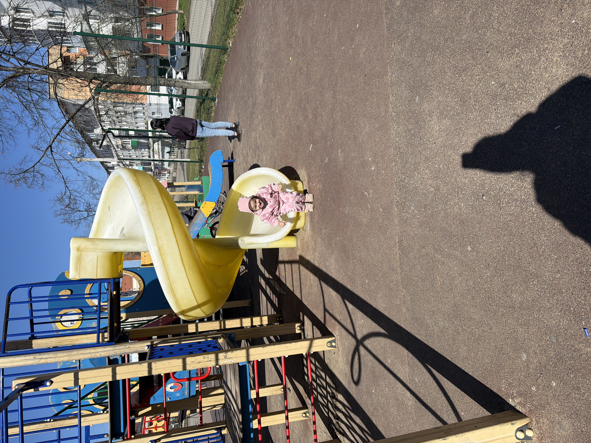 A child, dressed in a pink snowsuit, sits at the bottom of a yellow slide on a playground in Chaikovsky, Russia. A woman stands nearby. The playground is on a brown, textured surface. A multi-colored play structure with blue, yellow, and red elements is visible. A building is in the background, partially obscured by bare trees. A tall shadow is cast onto the playground surface.