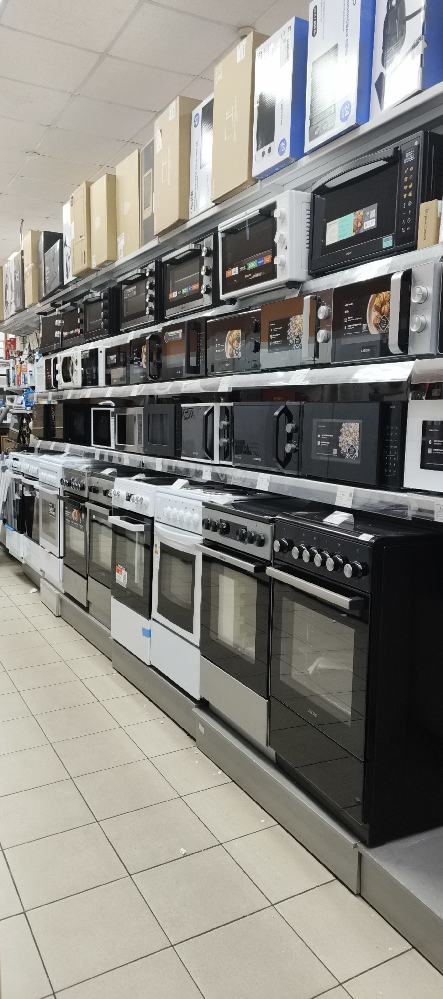 The image shows a retail store aisle filled with various kitchen appliances, primarily ovens and microwaves. The appliances are arranged on multiple shelves, with boxes of some items stacked above. The store appears to be well-stocked, showcasing a wide selection of these domestic appliances. The flooring is tiled in a neutral color. The image is taken from a slightly elevated perspective, looking down the aisle. No people are visible in the frame. The overall impression is of a typical appliance retail environment. Based on the context, this scene is located in the city of Kaltan, Russia.