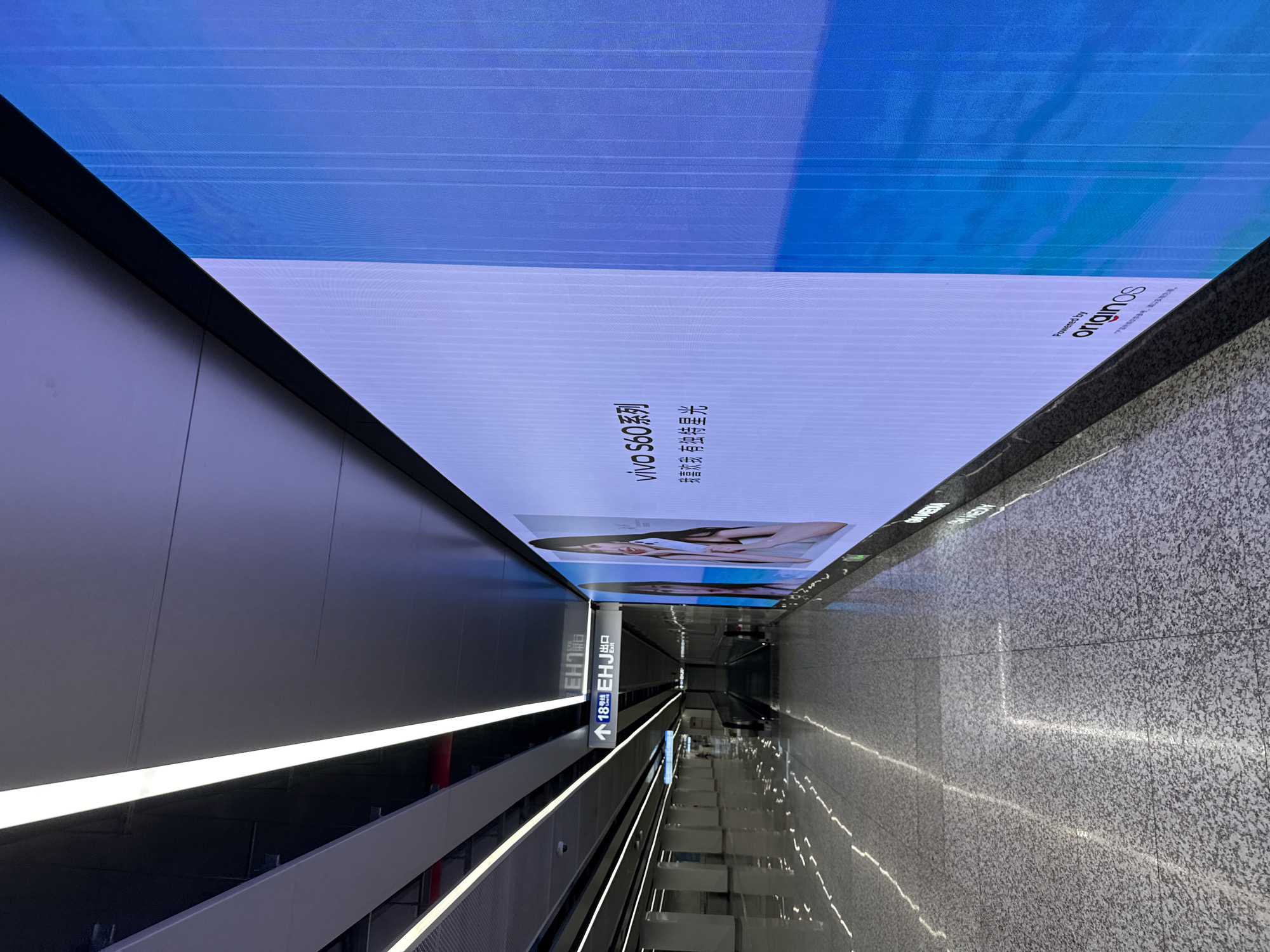 The image depicts a modern subway station in Haizhu District, China. A large digital billboard displays an advertisement for "vivo S60 series" with Chinese text. The billboard is illuminated and takes up a significant portion of the right side of the frame. The subway station features a clean, well-lit interior with polished floors reflecting the overhead lighting. On the left side of the image, a sign indicates "Line 18 EHJ Exit" with an arrow pointing forward, suggesting the direction of the subway line and an exit. An escalator is visible in the mid-ground. The overall environment is futuristic and orderly, typical of contemporary urban transit hubs.