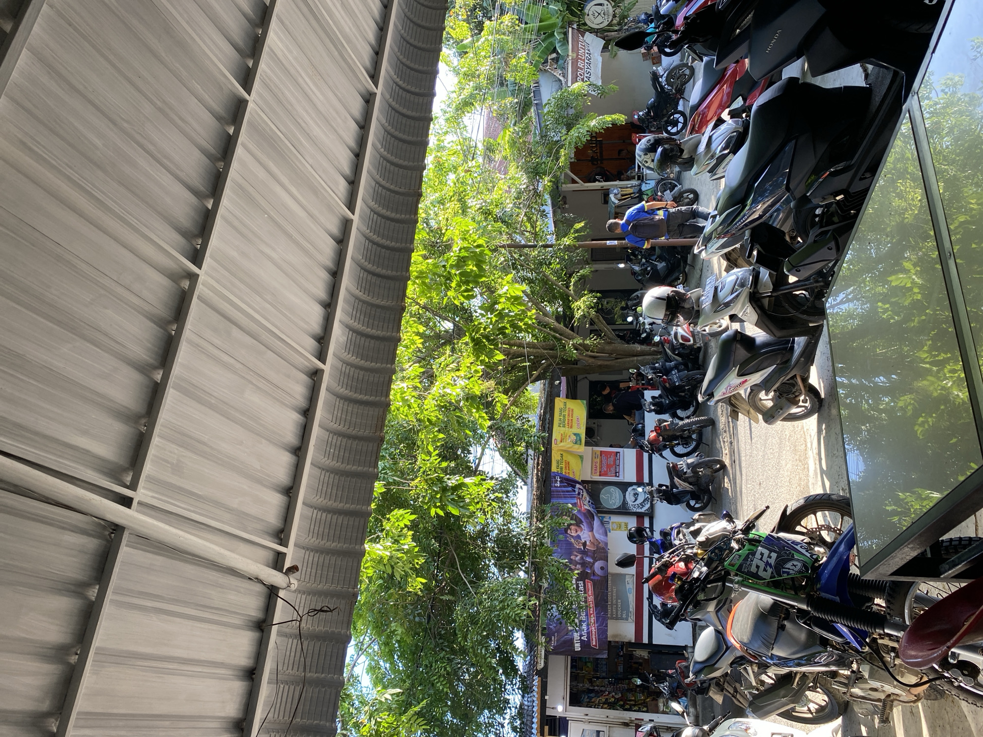 The image depicts an outdoor scene in Lhokseumawe, Indonesia, showcasing a parking area filled with motorcycles. The shot appears to be taken from under a corrugated metal roof structure, which frames the left side of the image. A dense canopy of green trees fills the central portion, partially obscuring storefronts and signage in the background. A variety of motorcycles, including models from Honda, are parked along the street, some angled towards the storefronts. Storefronts are visible behind the motorcycles. Signage indicates a variety of businesses including a pharmacy and businesses selling internet products. A blue sign displays the phrase "POLRI UNTUK ASYARAKAT" (Police for the Community). Another sign advertises "Morinaga Chiliga". One man is seen wearing a blue shirt and pants.
