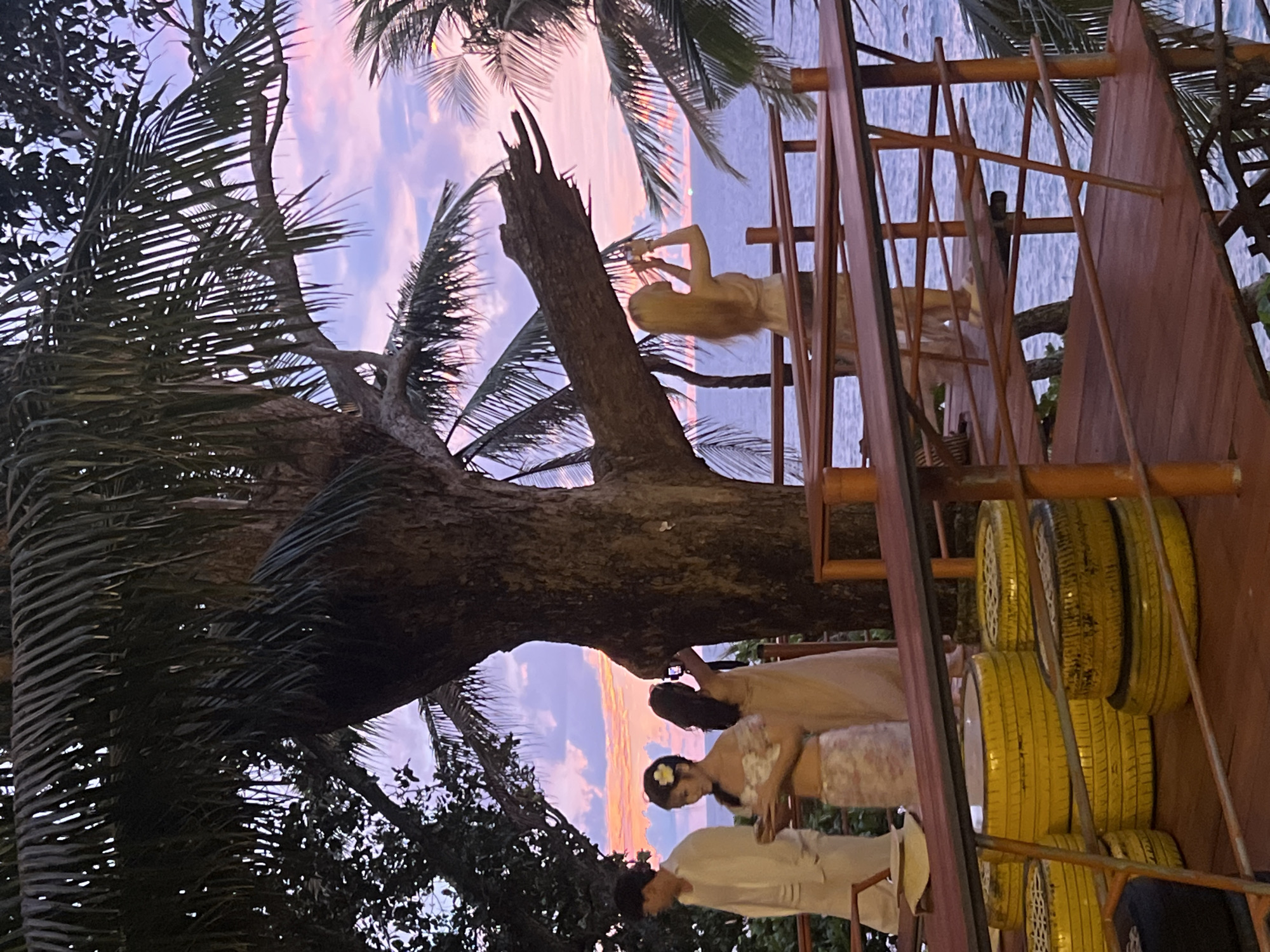 In Karon, Thailand, at dusk, a group of people are gathered on a wooden platform overlooking the sea. A man in a white suit stands on the left, his back to the camera. Next to him, a woman in a floral dress with a flower in her hair looks down at her hands. Behind her, another woman with dark hair is taking a photo with her phone. Further to the right, a woman with long blonde hair is also taking a photo with her phone, facing the ocean. The sky is filled with soft pink and purple hues from the sunset, with scattered clouds. Palm trees frame the scene, their fronds dark against the colorful sky. The wooden platform has a rustic appearance with railings made of bamboo and wood. In the foreground, stacked yellow tires are visible, some appearing to be used as makeshift seating. The ocean stretches out to the horizon, reflecting the soft light of the setting sun.