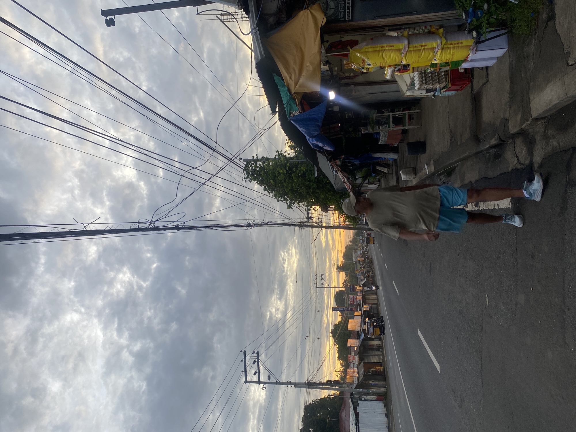 The image shows a man walking on the side of a road in what appears to be a commercial area at dusk. The man is wearing a light-colored t-shirt, blue shorts, and athletic shoes. He is walking away from the camera towards the right of the frame. The road is paved and has white lane markings. On the right side of the road, there are several small shops with corrugated metal roofs and awnings made of tarps. Some of the shops have goods displayed outside, including bags of rice and crates of eggs. There are power lines and utility poles running along the street. In the background, there are more shops and buildings, as well as some trees. The sky is cloudy with a warm glow suggesting either sunrise or sunset. The presence of what appear to be tricycles and motorcycles on the road indicates this is a typical street scene in the Philippines. Given the context of similar streetscapes, this location is likely within Davao City, Philippines.