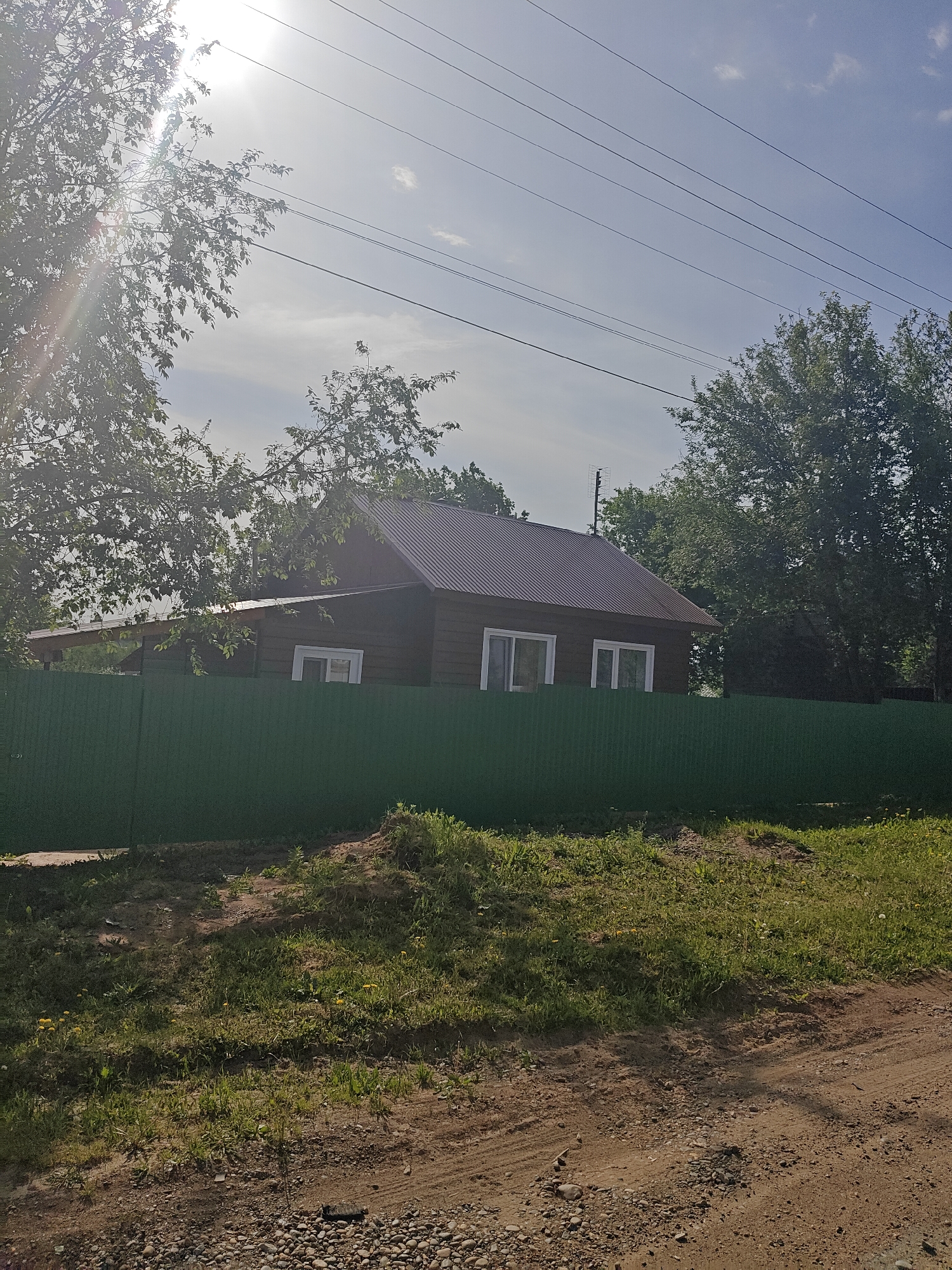 A daytime exterior shot captures a wooden house situated in Фоки, Russia, behind a green fence. The house has a brown roof, dark wooden walls, and white-framed windows. Tall green trees partially obscure the house on both sides. The ground in the foreground is a mix of dirt, gravel, and green grass. Power lines stretch across the upper part of the frame against a partly cloudy sky, with bright sunlight visible through the trees.