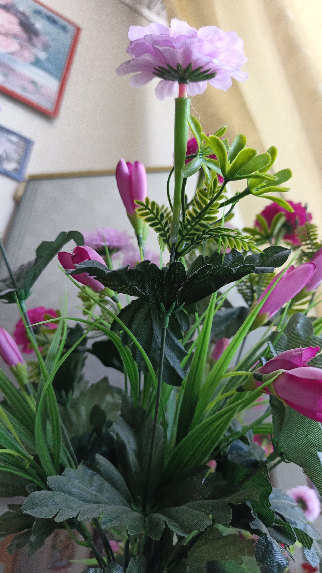 An upward-angle, close-up, eye-level shot focuses on a bouquet of artificial flowers. The bouquet is comprised of various pink and purple flowers, with abundant green foliage and stems. In the foreground, a large, dark green leaf with serrated edges dominates the lower portion of the frame. To the upper center, a light purple flower with delicate, layered petals is visible, attached to a green stem. Smaller pink, bud-like flowers are interspersed throughout the arrangement. The background is blurred, but appears to be an indoor setting with a light-colored wall and hints of framed pictures. The lighting is soft, casting a gentle glow on the flowers.