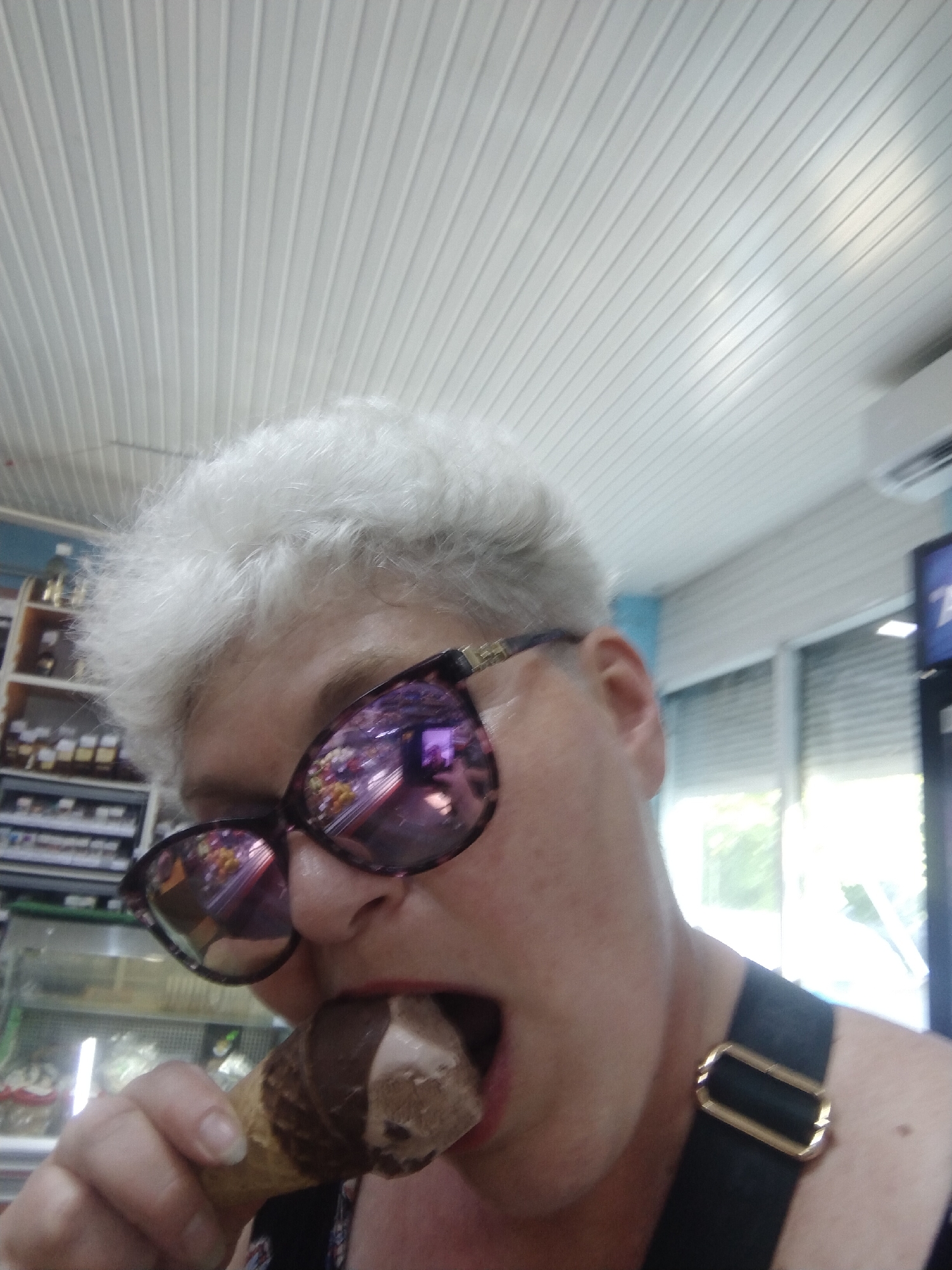 An older woman with short, white, curly hair is taking a selfie while eating an ice cream cone. She is wearing large, dark-rimmed sunglasses with pinkish lenses that reflect the surrounding environment, including what appears to be a retail store with shelves of products and a display case with food items. She is also wearing a black strap across her shoulder. The background shows a white, paneled ceiling, shelves stocked with various jars and bottles, and a window with blinds. The location is identified as Horishni Plavni, Ukraine.