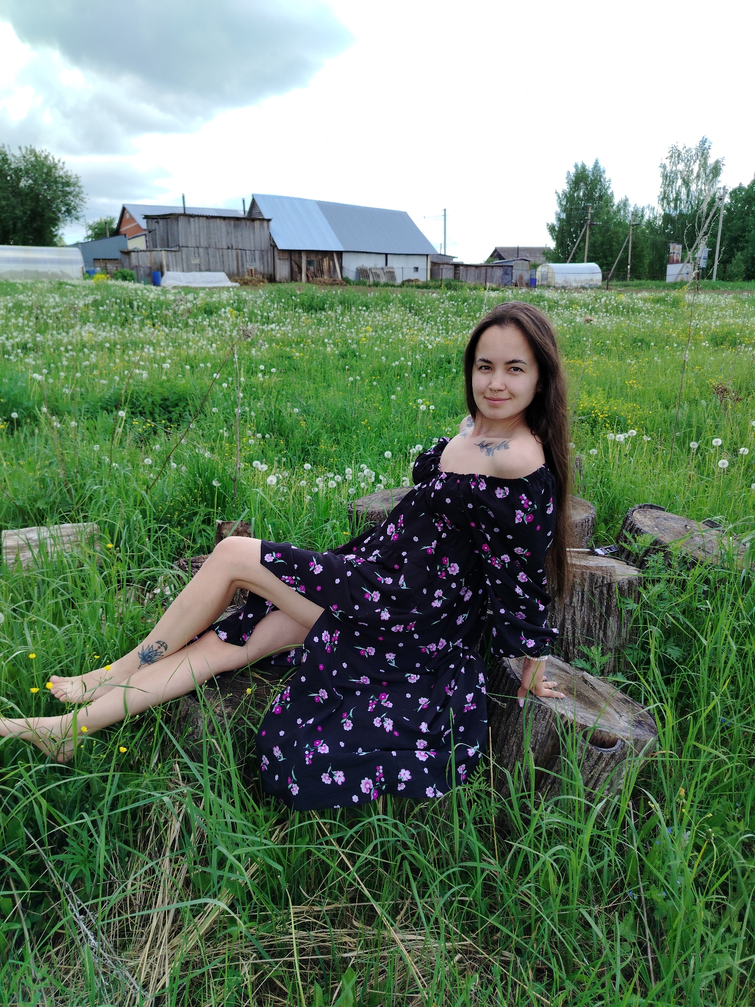 A woman sits on tree stumps in a grassy field in Гожан, Russia. She has long brown hair and is wearing a black dress with pink floral patterns. Her legs are bare and she has a tattoo on her ankle. In the background, there are wooden buildings and a field of wildflowers. The sky is overcast.