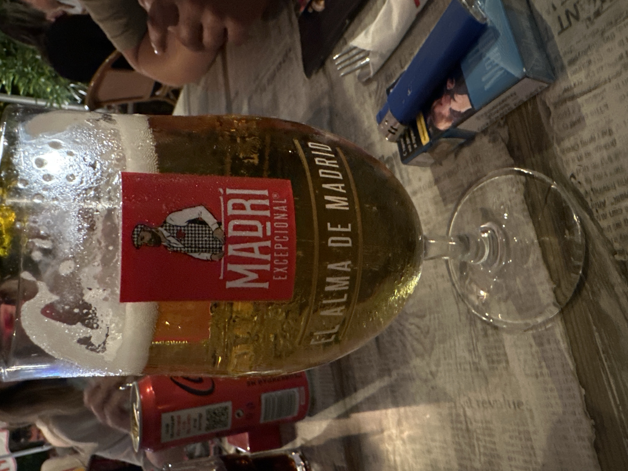 The image, rotated 90 degrees counter-clockwise, presents a close-up view of a table set with several drinks and personal items, likely in a bar or restaurant setting.
The central and dominant subject is a tall glass of beer, half-full with a golden liquid and a foamy head. Condensation beads are visible on the glass surface. A prominent red label on the glass reads "MADRI EXCEPCIONAL" and "EL ALMA DE MADRID" (The Soul of Madrid).
To the upper right of the beer glass, a blue cigarette lighter is placed on top of a blue cigarette packet. The packet displays a graphic of two individuals, consistent with health warning labels, and some unreadable text. A silver fork and a white folded napkin are also visible nearby. A clear, empty stemmed glass is partially seen to the far right.
In the lower left part of the frame, a red Coca-Cola can is present, showing its distinctive logo and a QR code. Adjacent to it, a small glass containing a dark-colored beverage is partially visible. The table surface is covered with a material resembling newspaper print, featuring columns of text, with the word "revolue" partially discernible in one section.
Parts of people are visible in the background and through reflections. In the top left corner, a person's arm, hand, and a portion of their head are visible, wearing a light-colored top. Reflections or outlines of another person can be seen in the lower part of the beer glass and near the Coca-Cola can. The lighting suggests an indoor or evening outdoor ambiance. No specific activity beyond casual drinking and socializing is evident.