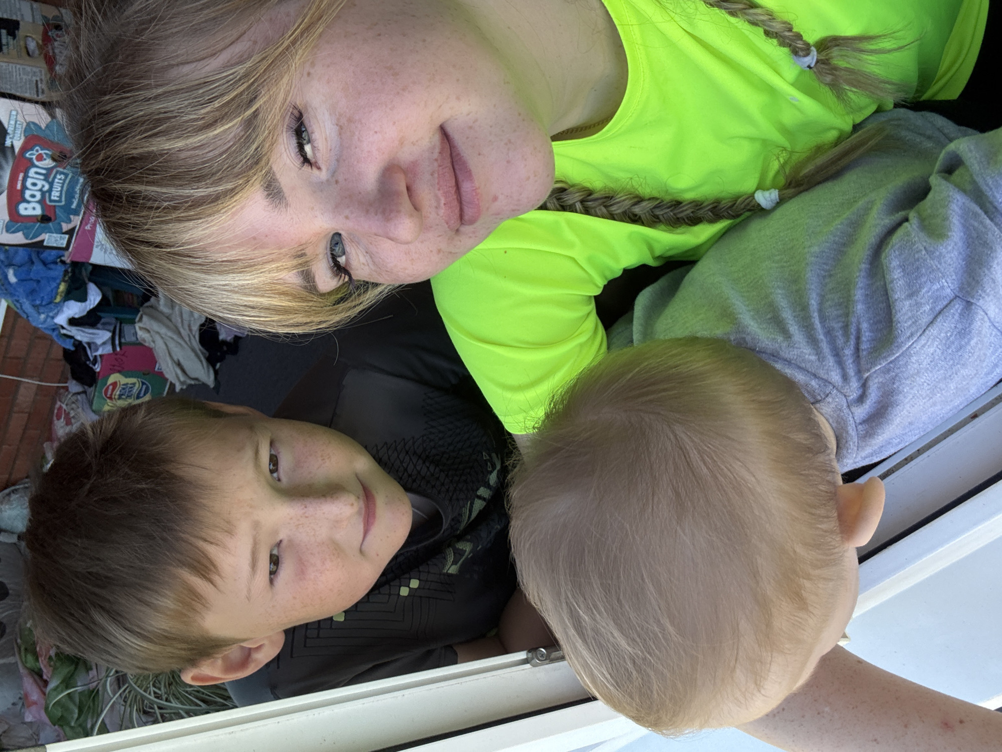 A selfie captures three individuals in an outdoor residential setting in Ляльшур, Russia. An adult female with blonde hair, bangs, two braids with white ties, blue eyes, and prominent facial freckles wears a neon green long-sleeved shirt, smiling faintly directly at the camera. To her left and slightly behind her, an adolescent male with brown hair and freckles wears a dark t-shirt with a geometric pattern, looking towards the camera with a subtle smile. In the immediate foreground, positioned below the female and to the right of the male, is an infant or toddler, visible from the top of its light blonde head and left ear, wearing a grey t-shirt and looking downwards. The individuals are clustered near a white window or door frame. The background is a cluttered outdoor space featuring a red brick wall, various items including stacked cardboard boxes (one labeled 'Bagn FRUITS' with an apple logo), a blue towel, other fabrics, and green foliage.