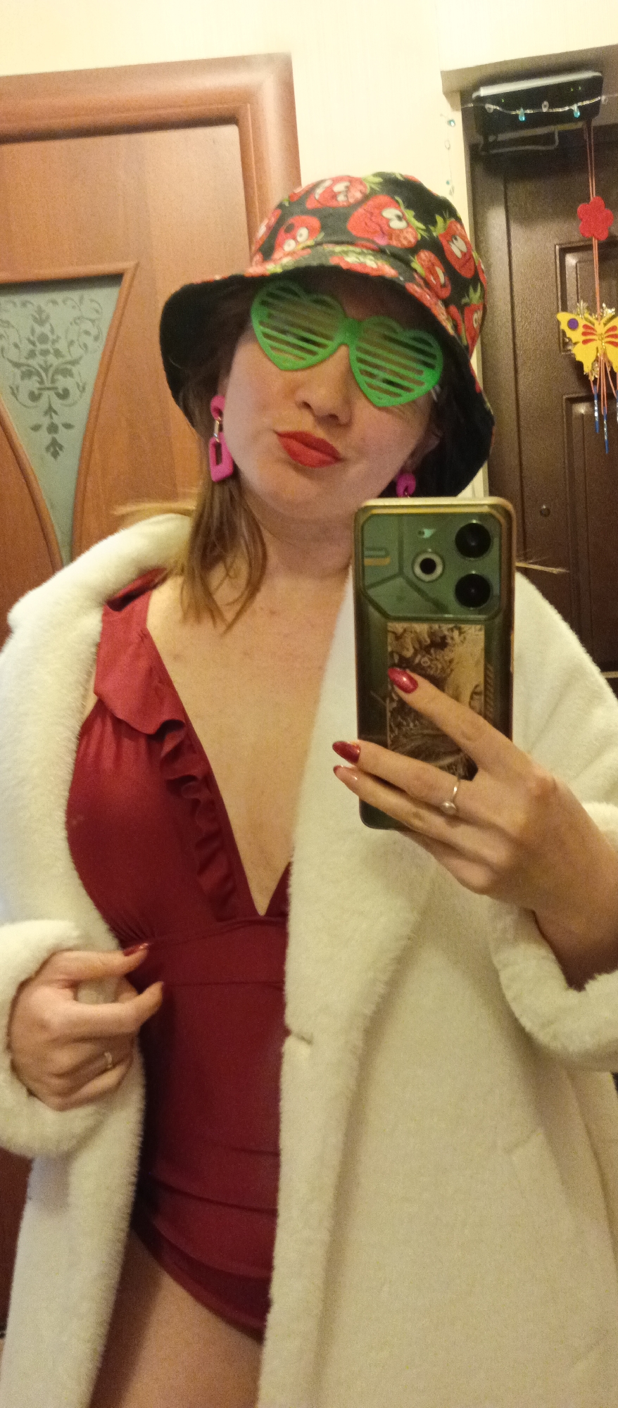 A medium close-up, eye-level selfie captures a woman indoors, posing for the camera. She is visible from the chest up, with her lips slightly pursed in what appears to be a playful or confident expression. She wears a black bucket hat adorned with a vibrant red and white strawberry pattern, and bright green heart-shaped shutter shades. Complementing her playful attire are striking pink rectangular dangle earrings and red lipstick.
She is dressed in a deep red swimsuit or low-cut top with a ruffled V-neckline, partially covered by a fluffy white faux fur coat or robe. Her right hand, featuring red polished nails, holds up a dark-colored smartphone with a distinctive camera module (two large lenses and one smaller lens) and a patterned case. A ring is visible on her left ring finger.
The background shows two light brown wooden doors. The door on the left has a decorative glass panel with an ornate pattern. The door on the right features a wall-mounted electronic device, possibly a router, and several colorful decorative hangings, including a yellow butterfly, a red flower, and a string of beads. The setting appears to be a casual home environment, possibly a hallway. No explicit text is clearly visible.