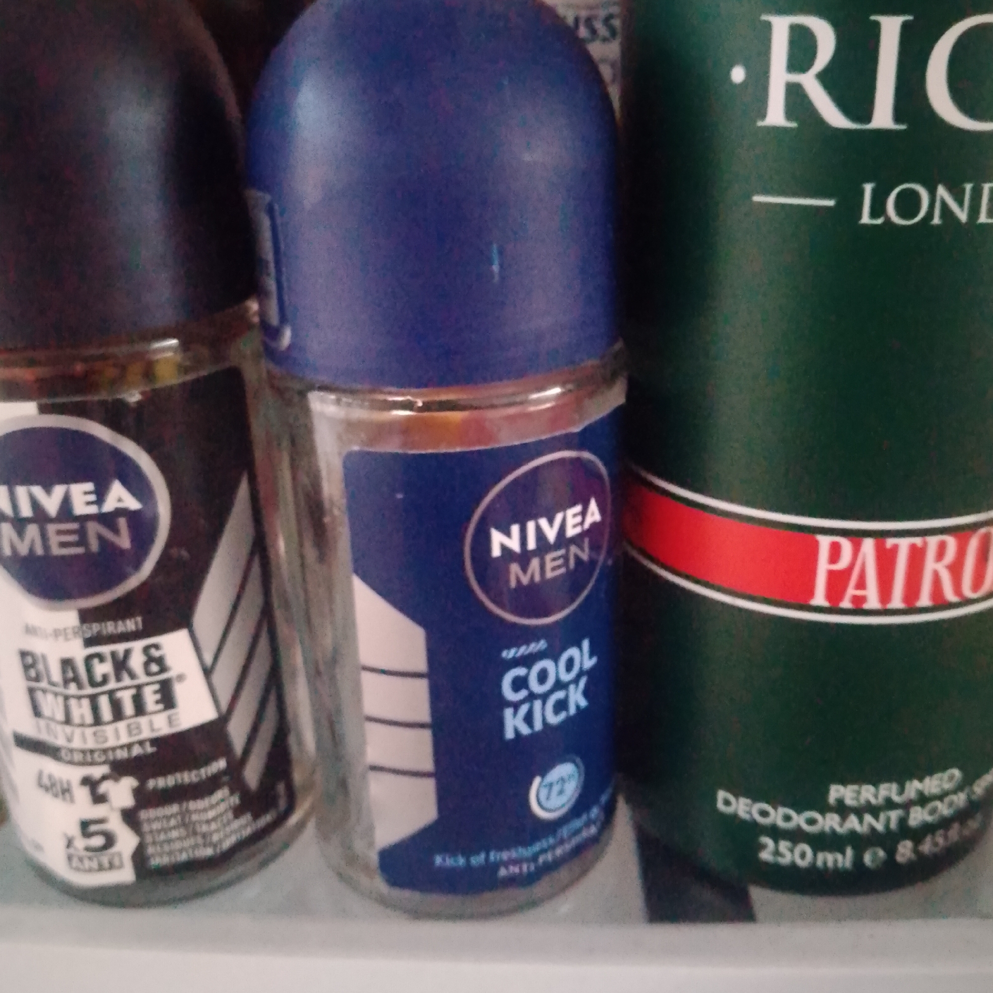 The image displays a close-up of three personal care products, likely antiperspirants or deodorants. On the left is a Nivea Men "Black & White Invisible Original" antiperspirant, featuring a black and white label with "48H" and "x5 ANTI-STAIN" text. Next to it is a Nivea Men "Cool Kick" antiperspirant with a blue label, showing "NIVEA MEN", "COOL KICK", and a "72h" indicator. To the right is a dark green cylindrical product, partially visible, with white text that reads "RIC", "LONDON", and "PATRO", followed by "PERFUMED DEODORANT BODY SPRAY 250ml". The products are arranged together, suggesting they are on a shelf or in a display. The lighting is even, indicating indoor conditions. No people or specific scene elements beyond the objects are visible. The text present on the products relates to their brand, function, and scent. No time of day, weather, or emotional cues can be inferred. The location context of Jalingo, Nigeria is provided but not visually evident in the image.