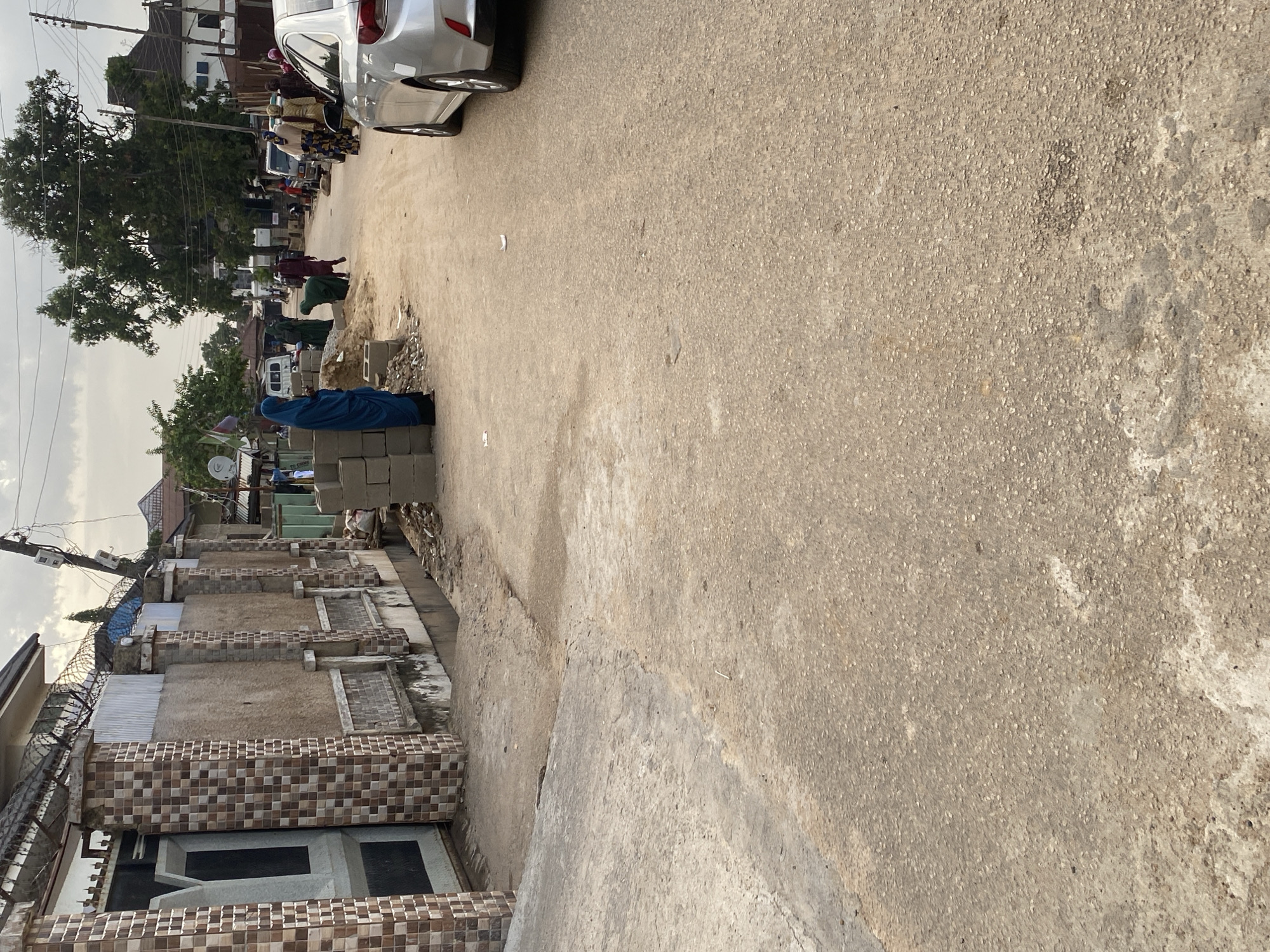 This is a street scene in Jos, Nigeria. The street is paved with asphalt and is lined with buildings and fences. In the foreground, a silver car is parked on the right side of the street. On the left side of the street, there is a fence made of concrete and tiles, with a large gate. Further down the street, several people are visible, some of whom are wearing traditional clothing. There are also stacks of cinder blocks and piles of dirt, suggesting ongoing construction or road work. The sky is overcast.