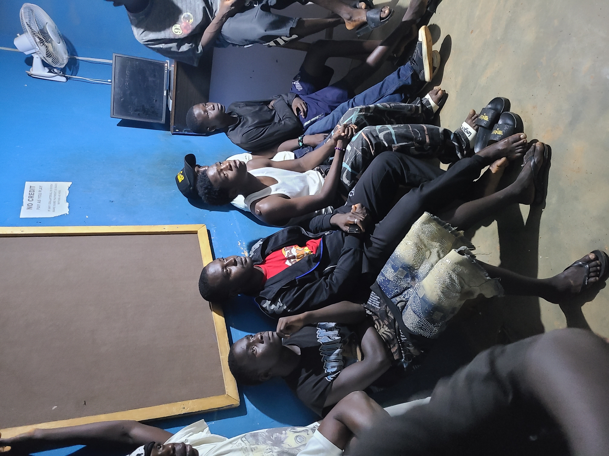 The image depicts a group of young men sitting in a dimly lit room with blue walls, possibly in Jos, Nigeria. The room appears to be a communal space, perhaps a gaming center or lounge. In the background, a computer monitor is visible on a shelf, along with a wall-mounted fan and a sign that reads "NO CREDIT PAY AS YOU PLAY". Several young men are seated on the floor and on a low platform, facing towards the right side of the image. They are dressed casually, with some wearing t-shirts and shorts, while others have on long pants. Some of the men are looking in the same direction, suggesting they are watching something or engaged in a shared activity. The overall atmosphere seems relaxed, and the lighting creates shadows that add to the subdued ambiance of the scene.