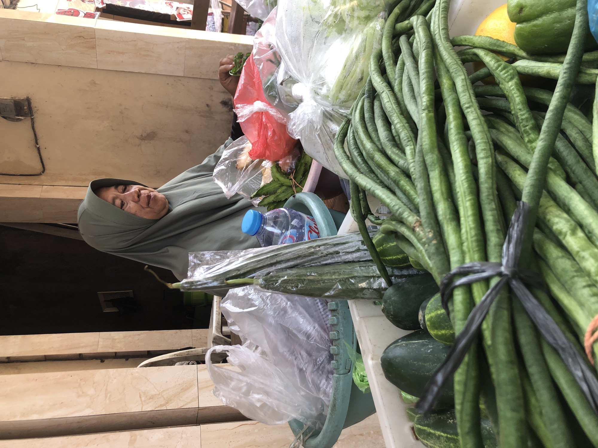 A woman wearing a sage-green hijab and a dark top is visible from the chest up, seated in front of a market stall. Her face, showing wrinkles and a mild expression, is turned to her right. Her right hand is holding a bunch of green leafy vegetables. The stall displays a variety of fresh produce in containers. In the foreground, a large bundle of long green beans is tied with a black plastic band. Behind the beans, a yellow lemon and a green bell pepper are partially visible. Other vegetables, including what appear to be cucumbers and possibly gourds, are arranged in plastic containers. A light blue bottle of water is also present on the stall. The background shows a building with beige tiled walls and a dark opening. The overall scene suggests an outdoor market setting in Surabaya, Indonesia, likely a traditional street-side vendor.