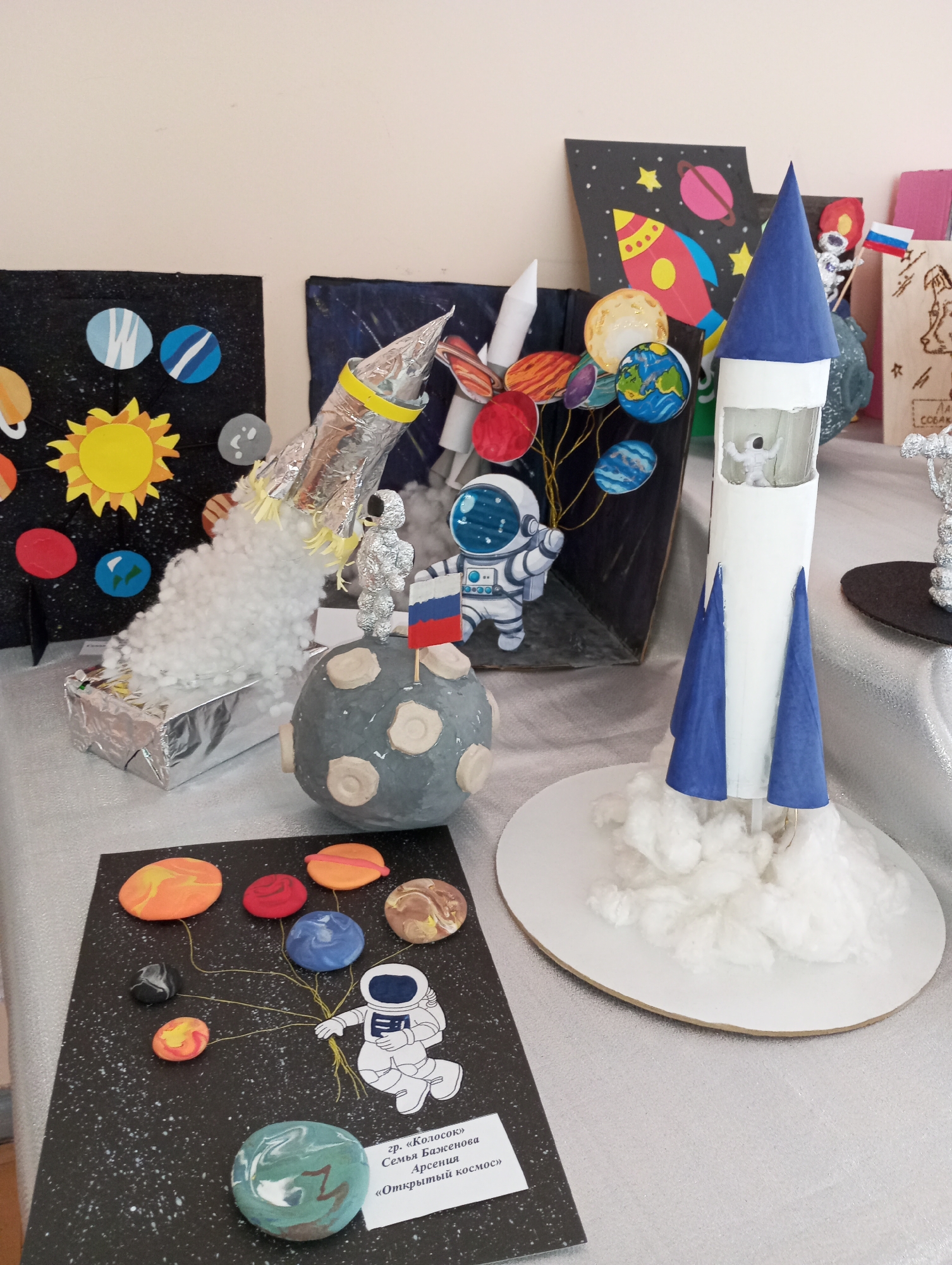 The image shows a display of student art projects related to space exploration. Several individual projects are visible, including representations of rockets, planets, and astronauts. One project features a cardboard rocket with blue fins and a cone-shaped top, positioned on a white base with cotton ball "smoke." Another features a rocket wrapped in foil. Cotton is used to represent smoke. A black rectangular board shows the sun and planets with the Earth. The projects sit on a metallic silver table. A written label is affixed to a planet-themed display in the lower left corner. The setting appears to be an indoor display or exhibition space.