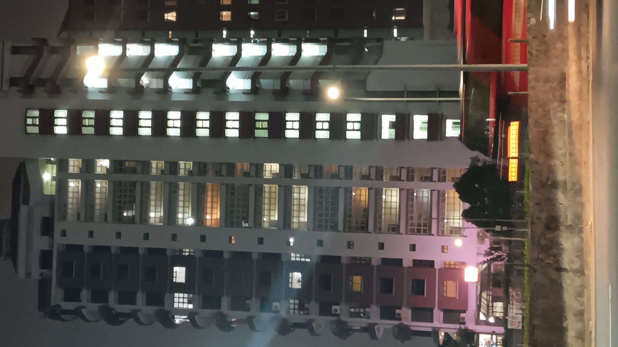 At night, a tall, modern apartment building stands illuminated by interior lights, casting a warm glow through its numerous windows. The building features a segmented facade with balconies and visible air conditioning units. To the right, another building, partially obscured, also shows illuminated windows. In the foreground, a red bus with illuminated yellow stripes is visible, moving from right to left. The scene is set against a dark, hazy sky, with streetlights casting a bright, ethereal light on the building's exterior. The location is the city of South Jakarta, Indonesia.