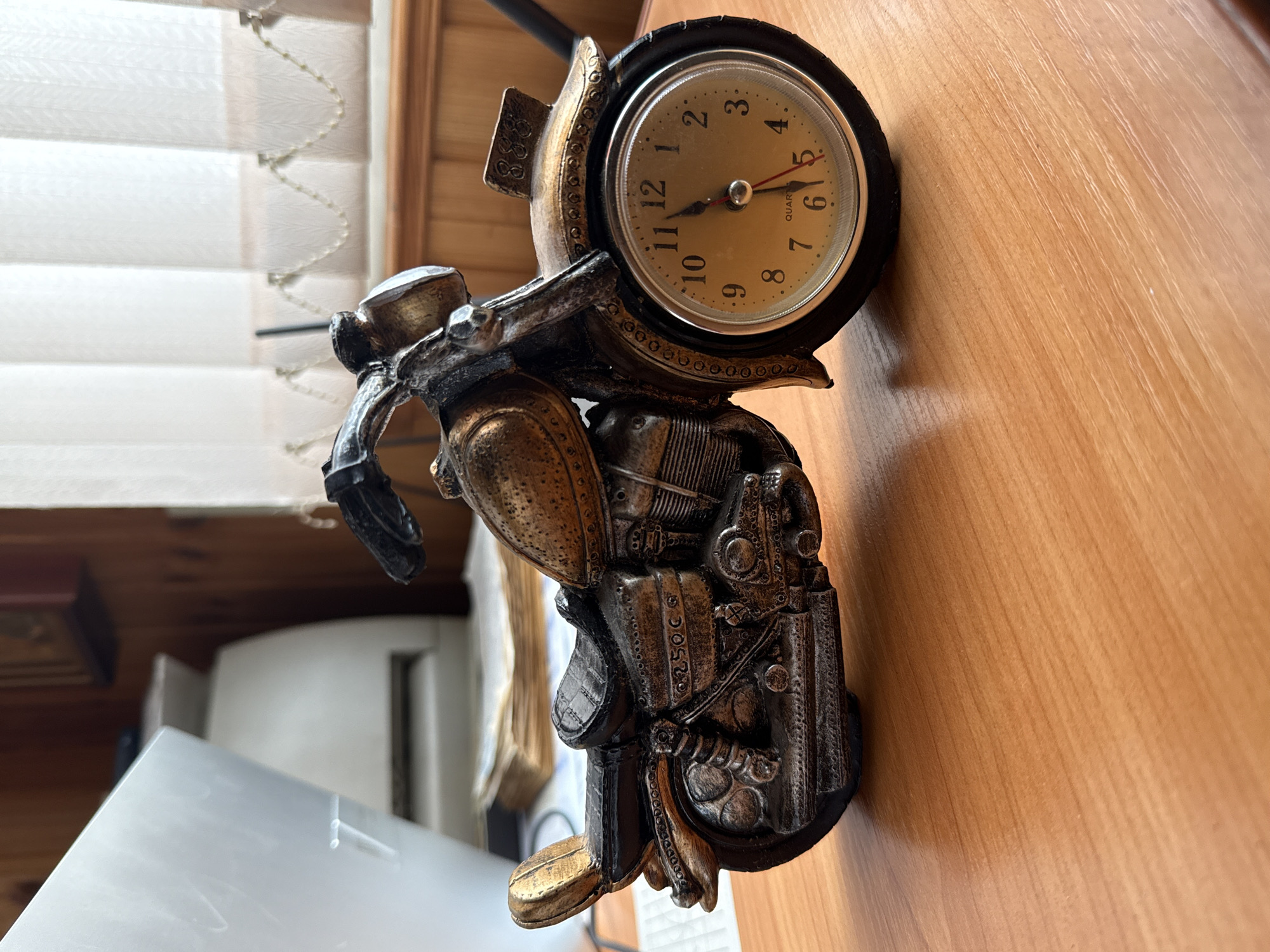 The image shows a decorative clock shaped like a motorcycle. The clock face is round with black numerals and hands. The motorcycle is intricately detailed, with a bronze and silver finish, and appears to be made of metal. The clock is positioned on a wooden surface, likely a desk, with a blurred background of blinds and wooden paneling. There is some visible text on the clock face which reads "QUARTZ" and some numbers. The overall lighting suggests it is daytime. No people are visible in the image.