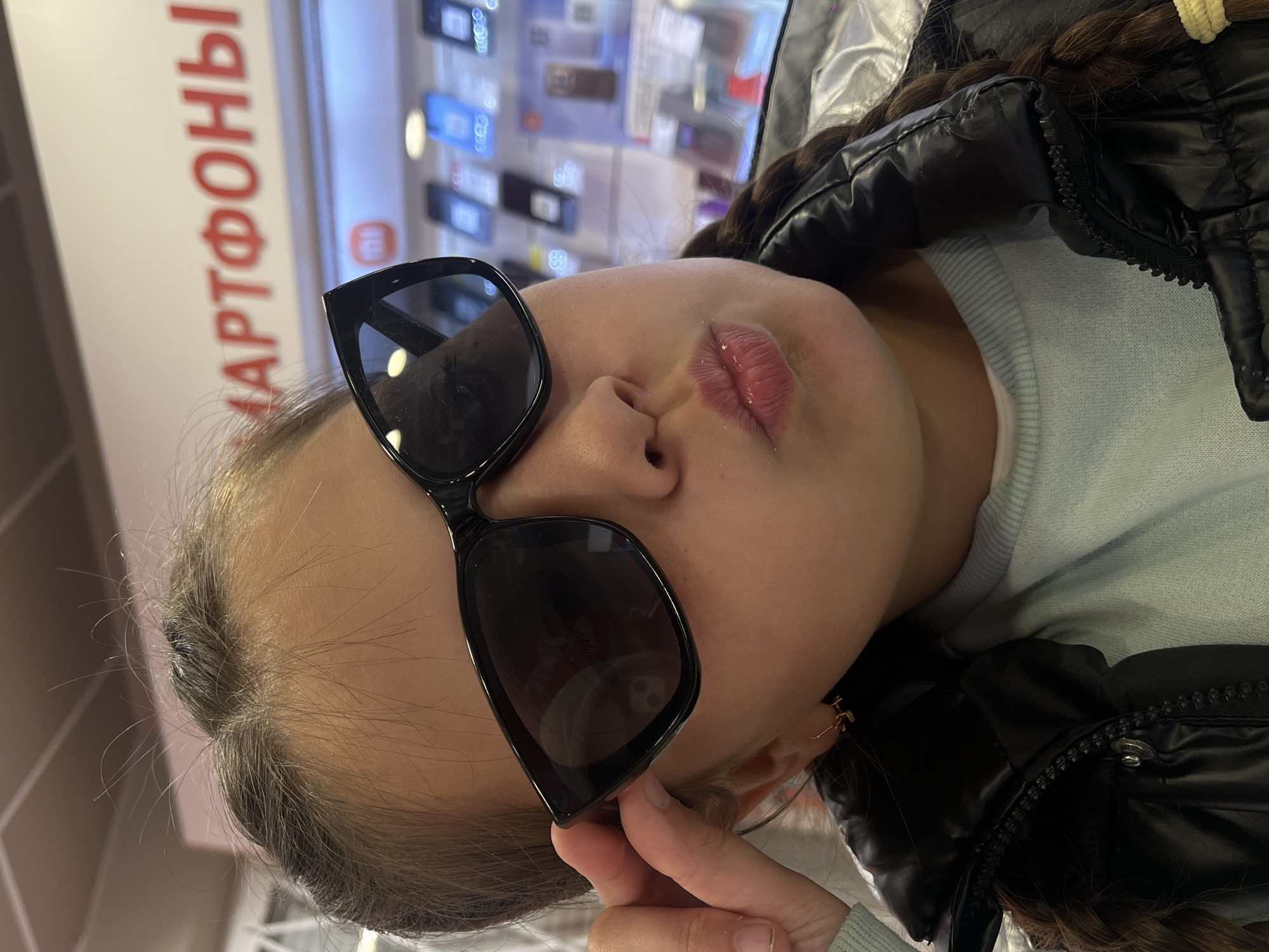 A close-up, slightly off-kilter portrait captures a young person, likely female, posing indoors. She is wearing large, dark sunglasses and has her lips subtly pursed, accented with glossy lip makeup. Her long, dark brown hair is visible, styled in a braid over her shoulder. She wears a black puffer jacket over a light blue long-sleeved top. Her right hand is lightly touching the frame of her sunglasses.
The background, which is out of focus, reveals display shelves showcasing multiple smartphones or electronic devices, suggesting the setting is an electronics retail store. A vertical sign with Cyrillic text is prominently visible in the background. The discernible part of the text reads "АРТФОНЫ", which is part of the Russian word "СМАРТФОНЫ" (SMARTPHONES), confirming the store's nature. The environment appears brightly lit. The image was taken in Novopokrovskaya, Russia.
