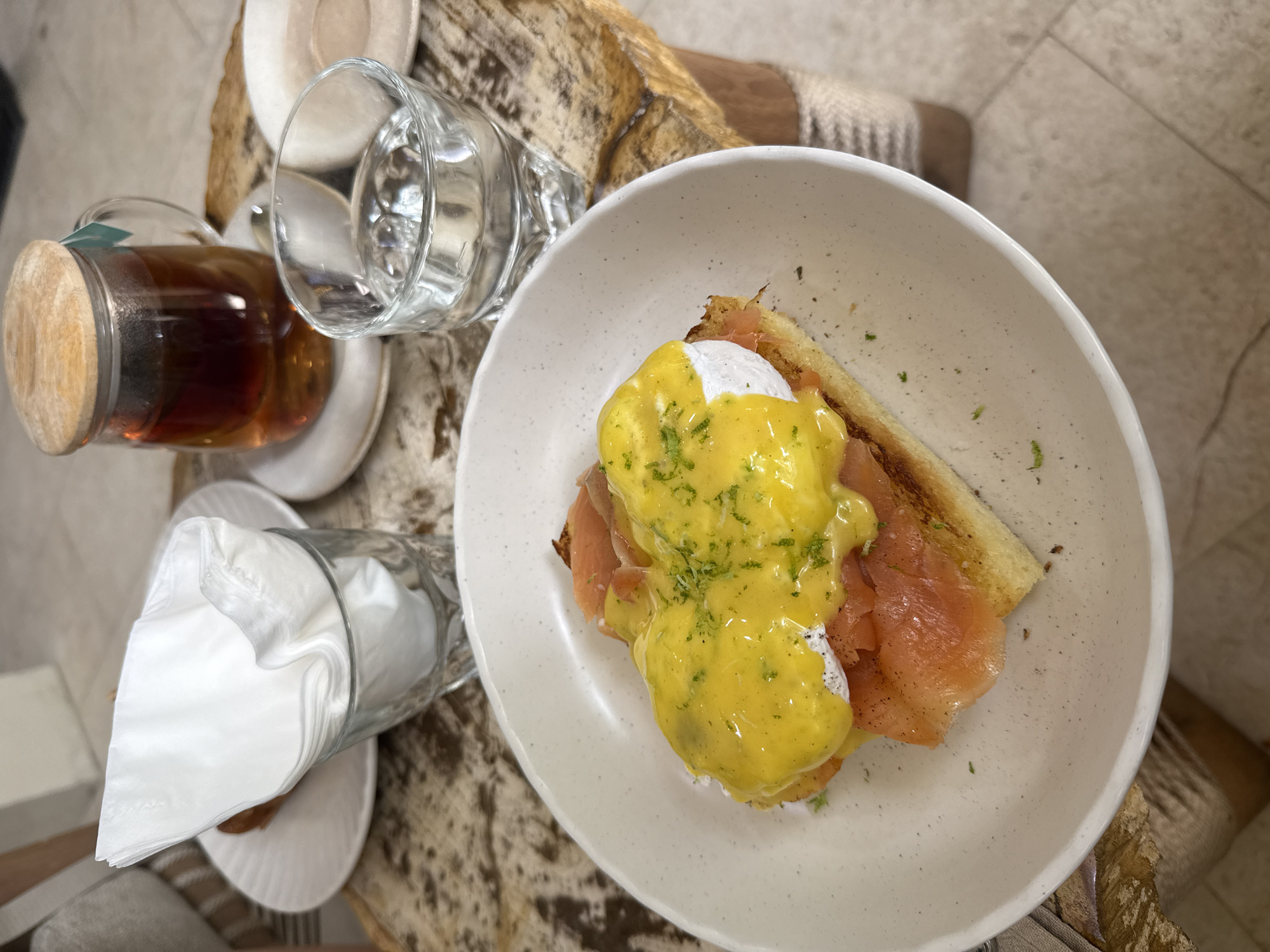 A high-angle shot captures a breakfast setting. In a white bowl, a serving of Eggs Benedict with smoked salmon on toast is presented. Beside the bowl, there's a glass of water and a tea infuser filled with dark liquid, accompanied by a glass containing white napkins. The scene is set on a textured surface of light and dark browns. The location is Mengwi, Indonesia.