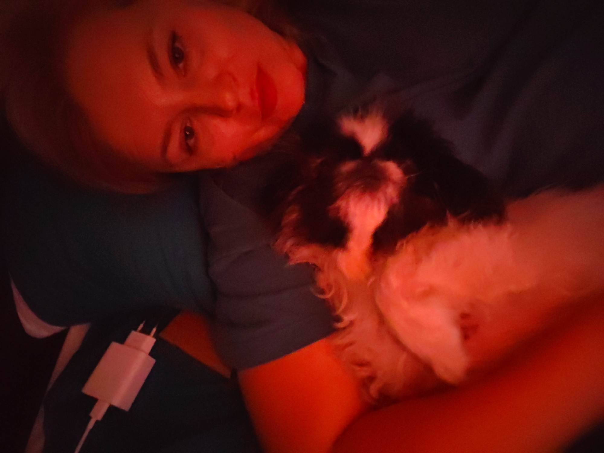 The image shows a close-up, dimly lit, overhead view of a person lying down and holding a small, fluffy dog. The person's face is partially visible on the left side of the frame, illuminated by a reddish-orange light, obscuring some of their features. Their eyes are open, and they appear to be looking at the camera. The dog is held close to the person's chest and is primarily white with black markings on its face and body. The lighting casts a strong reddish-orange glow over the entire scene, creating a warm and somewhat blurry atmosphere. The person is wearing a dark blue shirt or fabric, and a white electrical adapter with prongs is visible on their chest area, connected to an unseen cable. The background is dark and indistinct, offering no clear environmental cues. The image does not contain any identifiable landmarks or elements that would allow for its location to be specified in Izhevsk, Russia, or any other city.