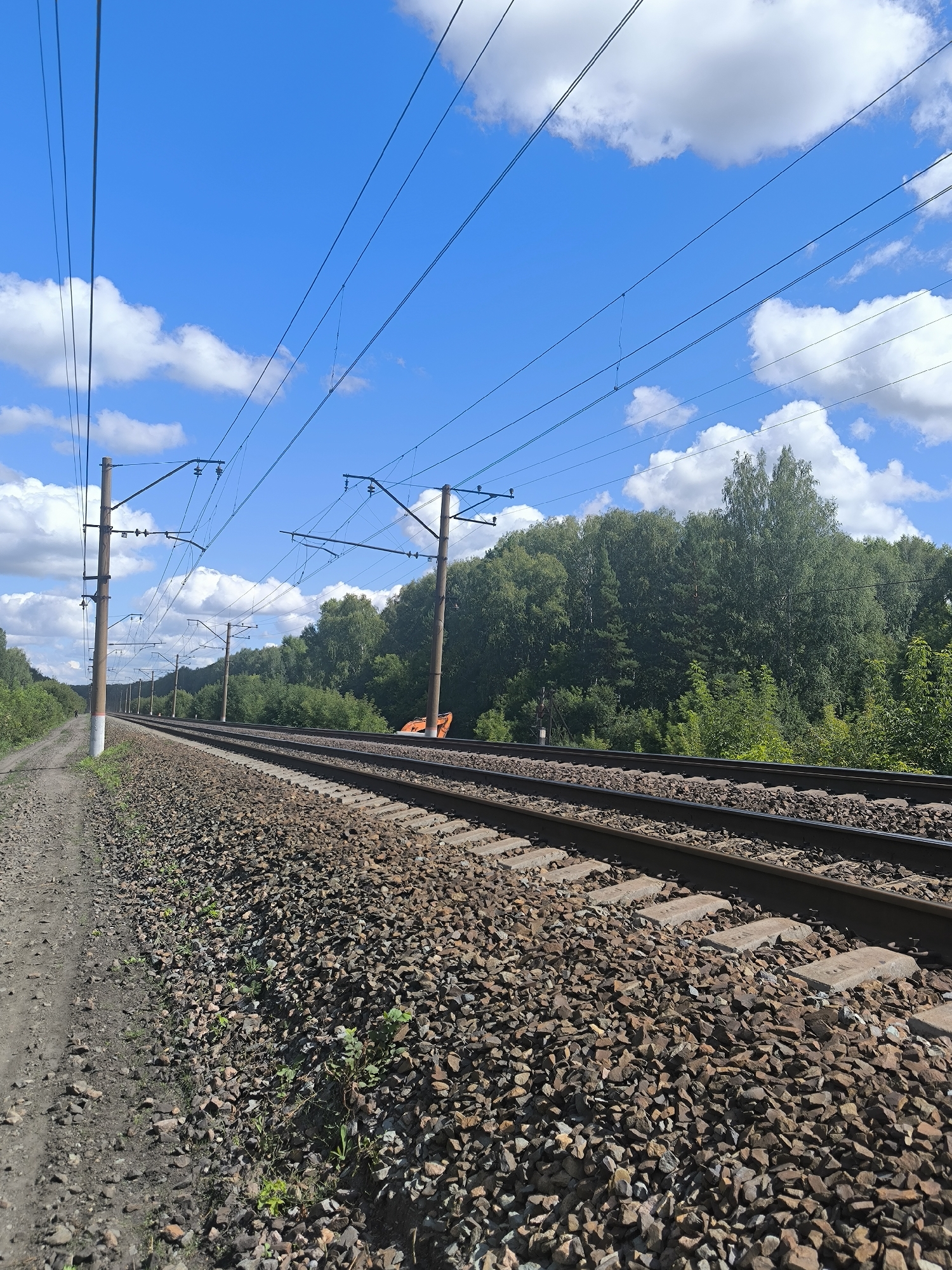 The image shows a railway track running parallel to a dirt path on the left. Overhead power lines are visible, supported by utility poles. The tracks are surrounded by gravel, and a dense forest lines the right side of the frame. The sky is blue with scattered white clouds. An orange excavator is partially visible among the trees to the right of the tracks. The scene is likely located near Maiskii, Russia, given the visual characteristics of the surrounding vegetation and infrastructure typical of the region. There are no people or animals visible in the image.