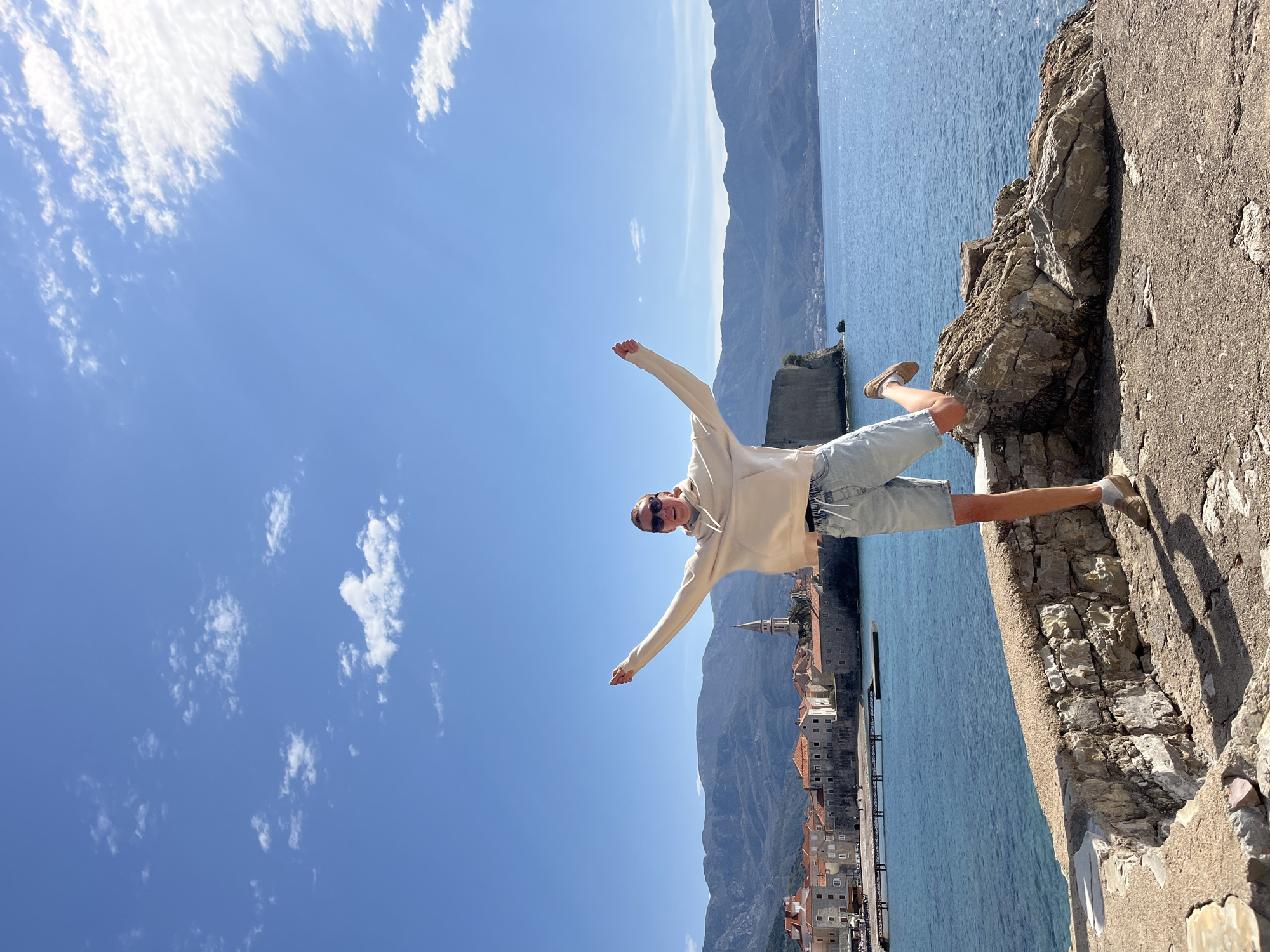 A person stands on a stone structure with their arms outstretched against a backdrop of clear blue sky and water. The person is wearing light-colored shorts, a light-colored hoodie, and shoes. The stone structure appears to be part of a coastal fortification or wall. In the background, there are mountains and a coastal town with red-tiled roofs. The town is part of Budva, Montenegro, as indicated by the architectural style and geographic location.