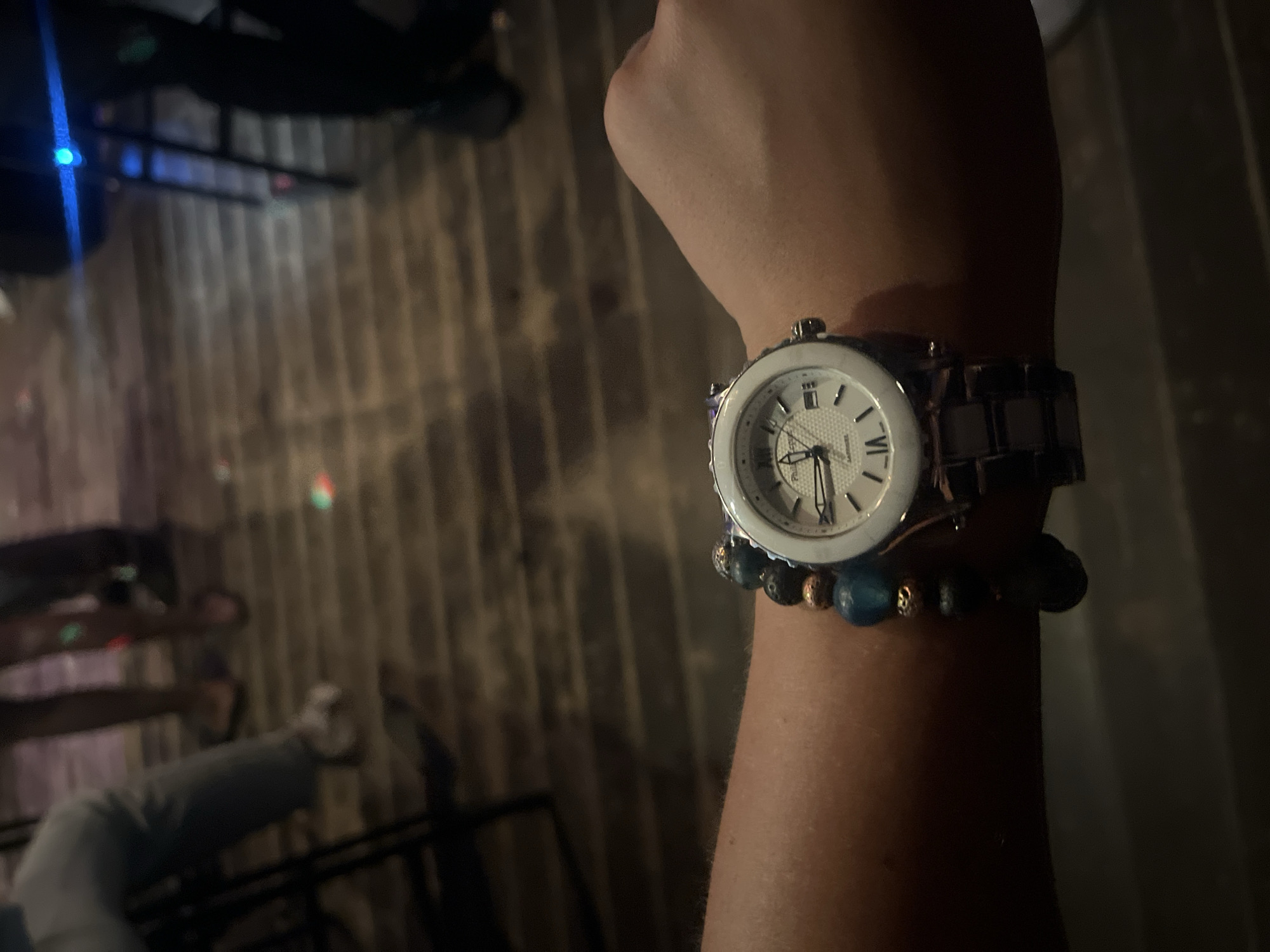 The image shows a close-up of a person's wrist wearing a metallic wristwatch with a white dial and a beaded bracelet. The background is out of focus, showing a wooden floor and the legs of people, suggesting an indoor social setting like a bar or restaurant. The lighting appears dim, with some colored lights reflecting on the floor. There is no discernible information about the location being in Pionersky, Russia.