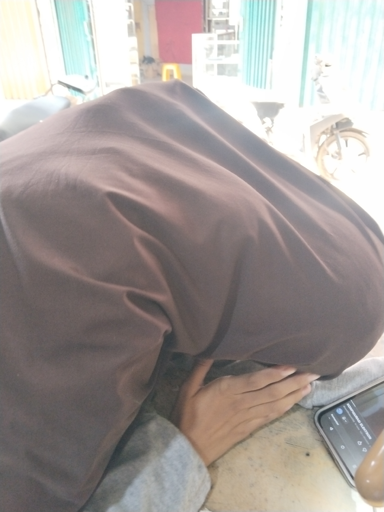 In the city of Unknown, Indonesia, a person is shown from a high angle, their head and upper body covered by a brown garment. A hand, appearing to be of an adult, is visible on the right side of the frame, resting on a gray sleeve. The hand is positioned near a smartphone, which displays text in Indonesian, including "KONFIGURASI ELEKTRON". The phone is lying on a wooden surface, likely a table. In the background, out of focus, are elements of an urban environment, including what appears to be a blue corrugated metal building, a yellow stool, and a motorcycle. The overall lighting suggests an outdoor setting with bright sunlight.