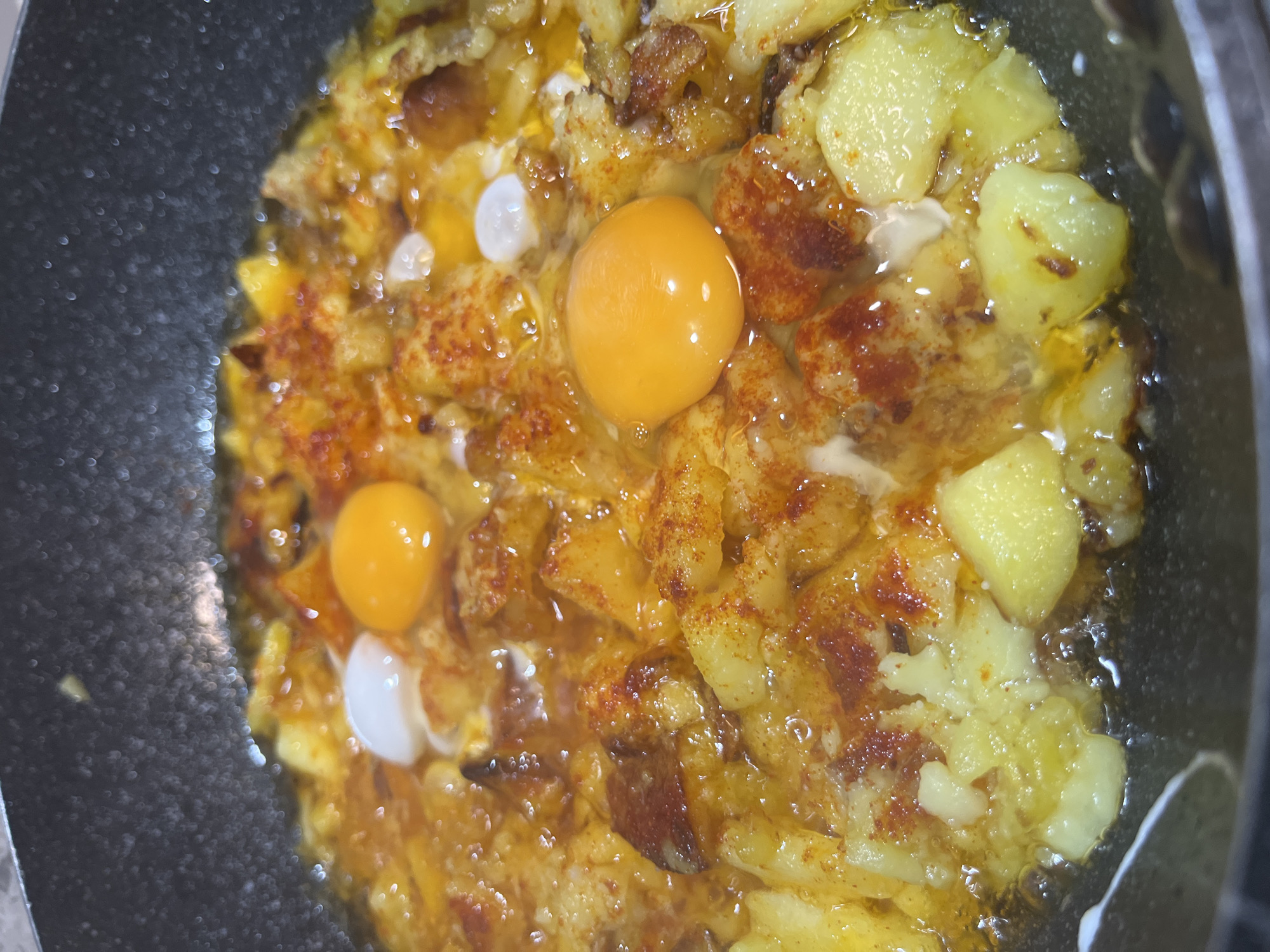 A close-up, top-down view captures a pan of fried potatoes and eggs, seasoned with paprika. Two whole eggs, one with a visible yolk and the other with a partially cooked white, are nestled amongst chunks of golden-fried potatoes. The potatoes are unevenly cut and appear to have some darker, crisped edges. A reddish-orange spice, likely paprika, is sprinkled generously over the dish. The food is in a dark, non-stick frying pan with a textured surface. The background is blurred, but the metallic sheen of the pan is visible. The scene is indoors, likely a kitchen. There is no indication of the specific location in Varna, Bulgaria.