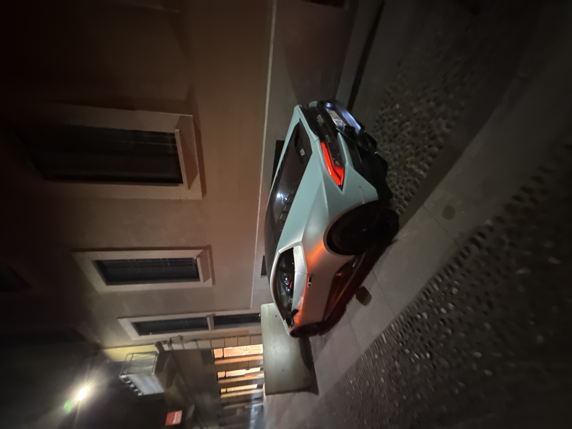 The image displays a modern, light-colored car, possibly a sports car or sleek coupe, parked on a street in Milan, Italy, at night. The car, featuring a contemporary design with sharp lines, has its red taillights illuminated and an orange glow reflecting on its front right side, possibly from a turn signal or ambient light. Interior dashboard lights are also visible within the cabin.
The vehicle is positioned alongside a building with a light-colored facade. This building features several recessed, rectangular windows, some of which appear to have grilles or horizontal blinds. The ground consists of a dark, irregularly shaped cobblestone pavement, transitioning to a smoother section closer to the building.
The scene is predominantly dark, illuminated by artificial light sources that highlight the car's reflective surfaces and parts of the building. The low light conditions emphasize the illuminated elements of the car and the textures of the street and building. No people are visible, and the primary activity is the car being parked. A license plate is present on the rear of the vehicle but is unreadable due to blurriness. The architecture and street paving are consistent with an urban European setting.