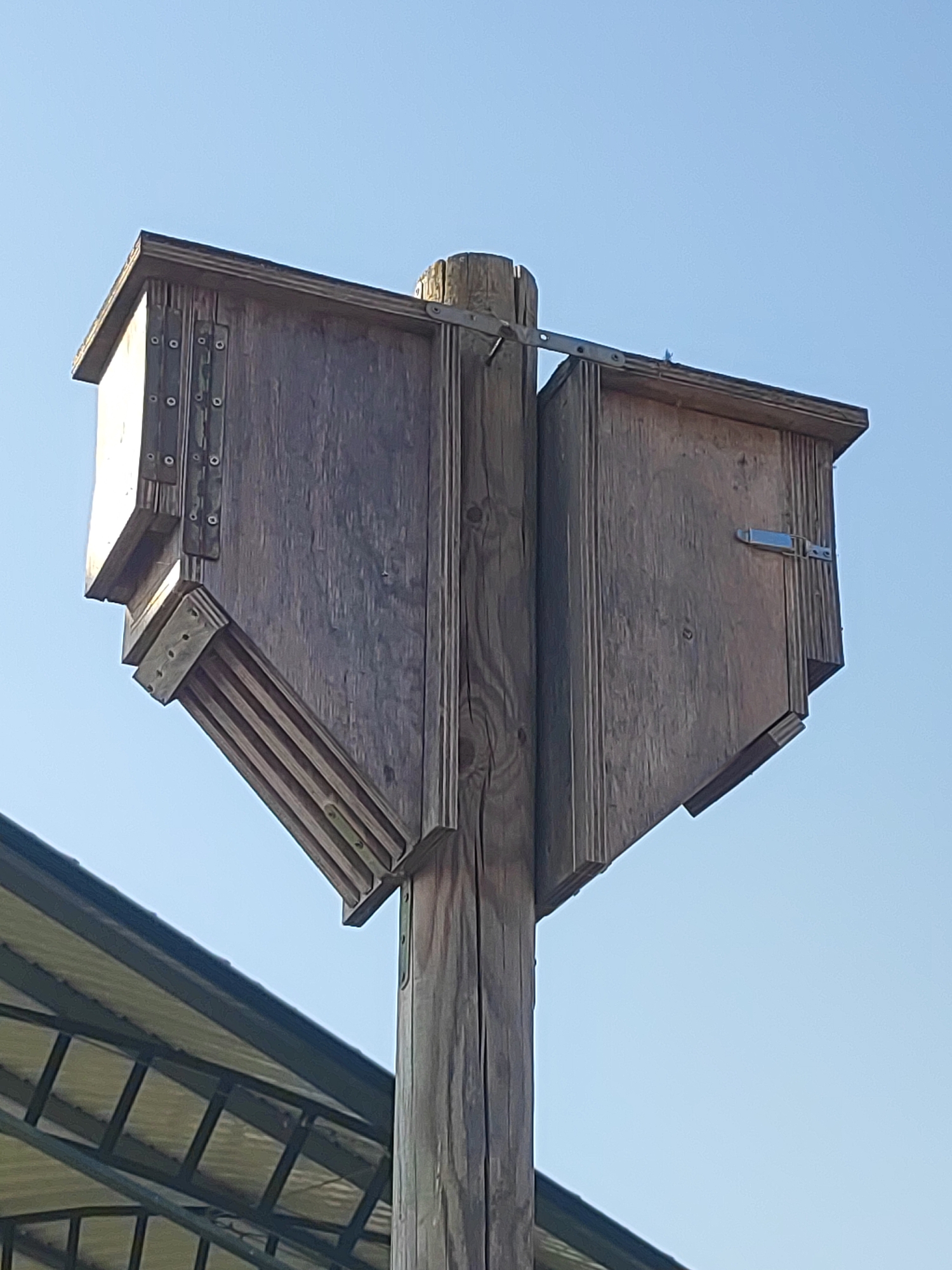 The image shows a wooden birdhouse mounted on a wooden post against a clear blue sky. The birdhouse is constructed of weathered wood and appears to have multiple compartments or entrances. One side of the birdhouse has a hinged metal latch. The birdhouse is positioned outdoors, and a portion of a metal-roofed structure is visible in the lower left corner of the frame. The scene is likely located in Autillo de Campos, Spain, given the context provided. There are no people or animals visible in the image.