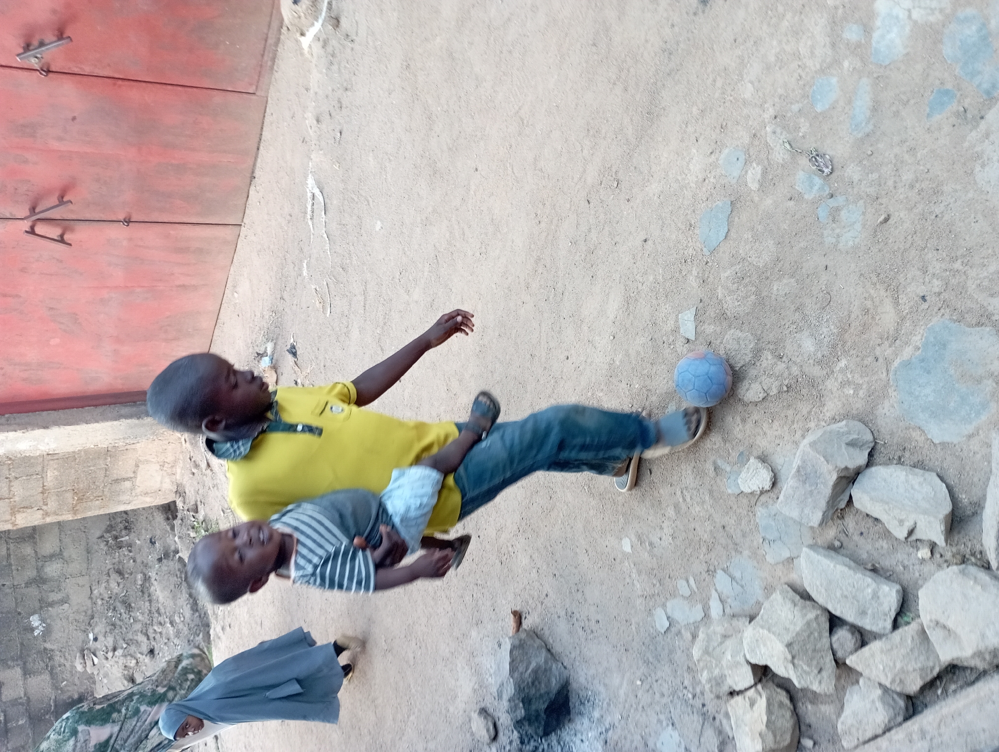 Two children, one in a yellow shirt and jeans holding another in a gray striped shirt and white shorts, stand on a dirt surface with scattered rocks near a blue soccer ball. A person in a blue robe stands in the background. The setting appears to be an outdoor area with a red metal door on the left and a partially visible building behind them. There are insufficient details to positively identify the specific location in Jos, Nigeria.