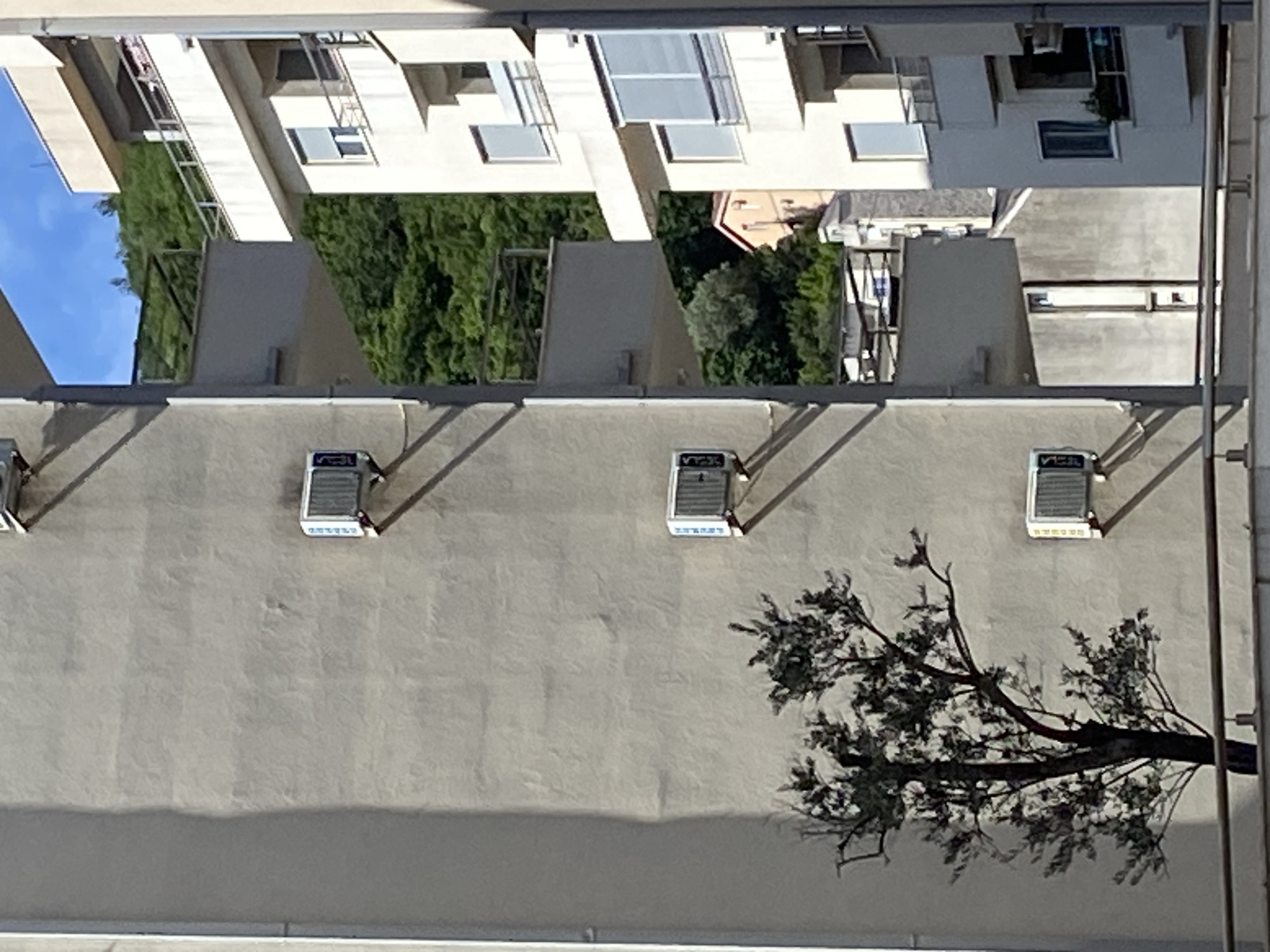 The image depicts an upside-down view of a building facade in Budva, Montenegro. The facade is gray with three air conditioning units mounted on the wall. A tree branch extends across the lower right portion of the wall. Above the facade, sections of buildings and vegetation can be seen, creating an inverted cityscape. The sky is visible in the upper left corner. Metallic bars run vertically along the side of the scene.