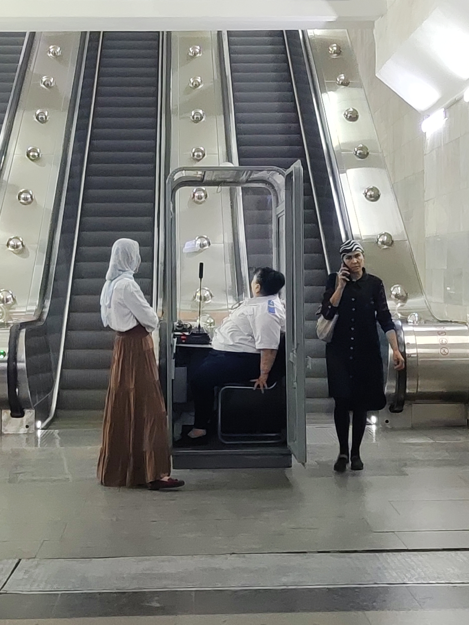 The image depicts a scene inside a subway station in Tashkent, Uzbekistan, specifically on an escalator platform. Three individuals are present. On the left, a woman wearing a light blue hijab and a long brown skirt stands facing away from the camera, with her arms crossed. In the center, a person in a white uniform with dark pants sits inside a glass-enclosed booth, appearing to be an attendant or operator. They are looking forward. On the right, a woman wearing a black, button-down coat and a black and white striped headscarf is on her phone and walking towards the right of the frame. Two escalators, with their characteristic ribbed black steps and polished metal sides adorned with circular decorative elements, dominate the background. The environment is brightly lit, with a tiled wall visible in the upper right corner. The flooring appears to be made of large, grey tiles.