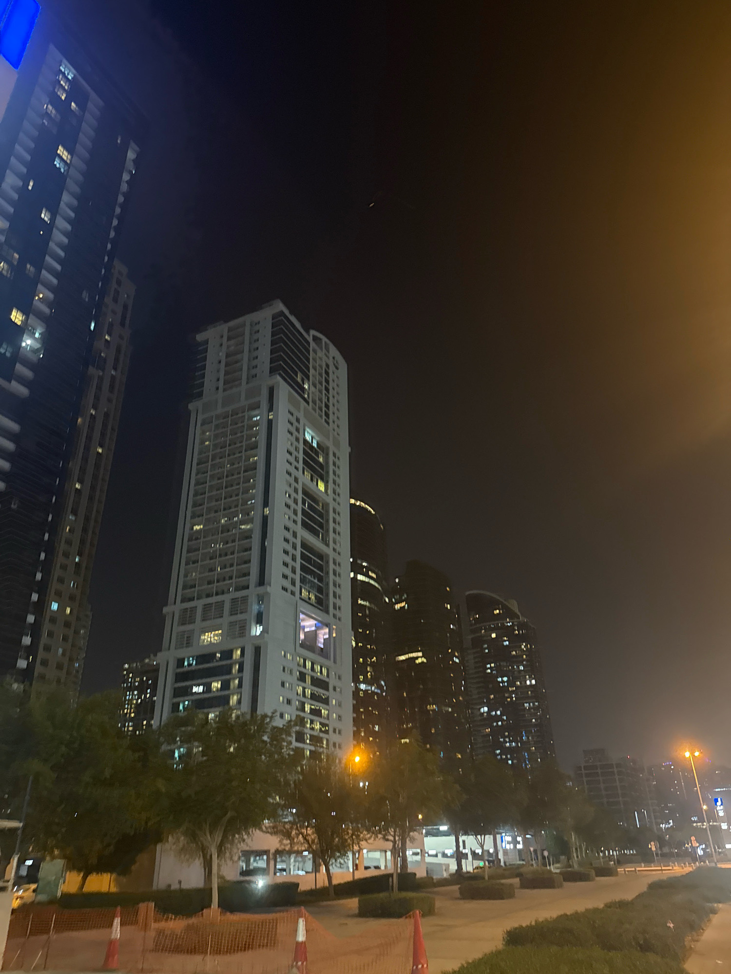 This is a night-time, low-angle, wide shot of a cityscape in Dubai, United Arab Emirates, featuring several modern high-rise buildings illuminated by interior lights and streetlights. The image is dominated by the dark sky and the glowing windows of the skyscrapers, creating a contrast between the urban development and the night.
In the foreground, there is a pathway lined with green trees and bushes, with some visible construction barriers with orange netting and traffic cones. The buildings are varied in design and height, with the most prominent being a tall, slender white skyscraper in the center, featuring a grid-like pattern of windows. To its left, another taller, darker skyscraper with a more uniform facade of windows is partially visible. Further in the background, a cluster of buildings with many illuminated windows suggests a densely populated urban area. A faint orange glow from streetlights or building lights colors the right side of the sky, indicating haze or smog. No people are clearly visible in the image.